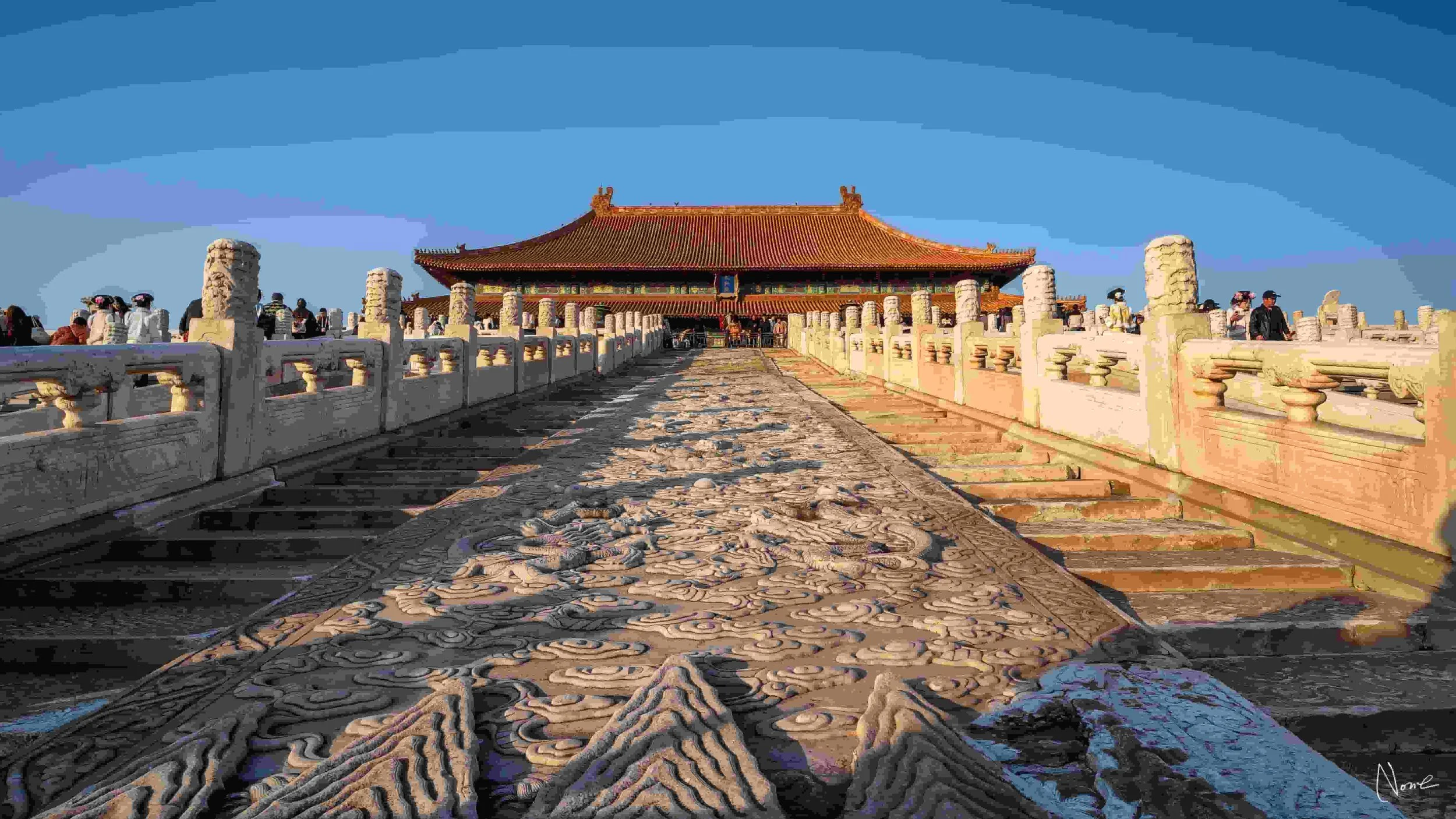 View of traditional Chinese palace buildings with orange-tiled roofs, surrounded by a red wall, under a blue sky with scattered clouds.