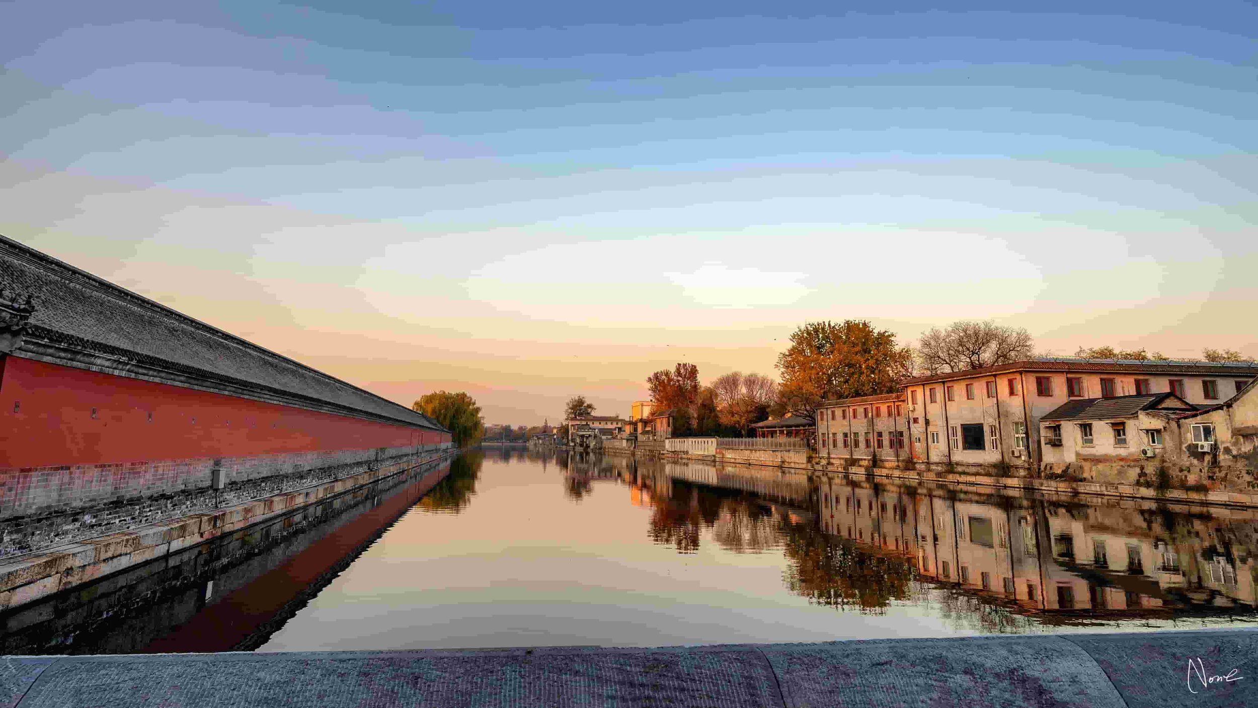 A peaceful canal scene at sunset with calm reflecting water, traditional Chinese architecture on the left, and modern buildings on the right, surrounded by trees with fall foliage.