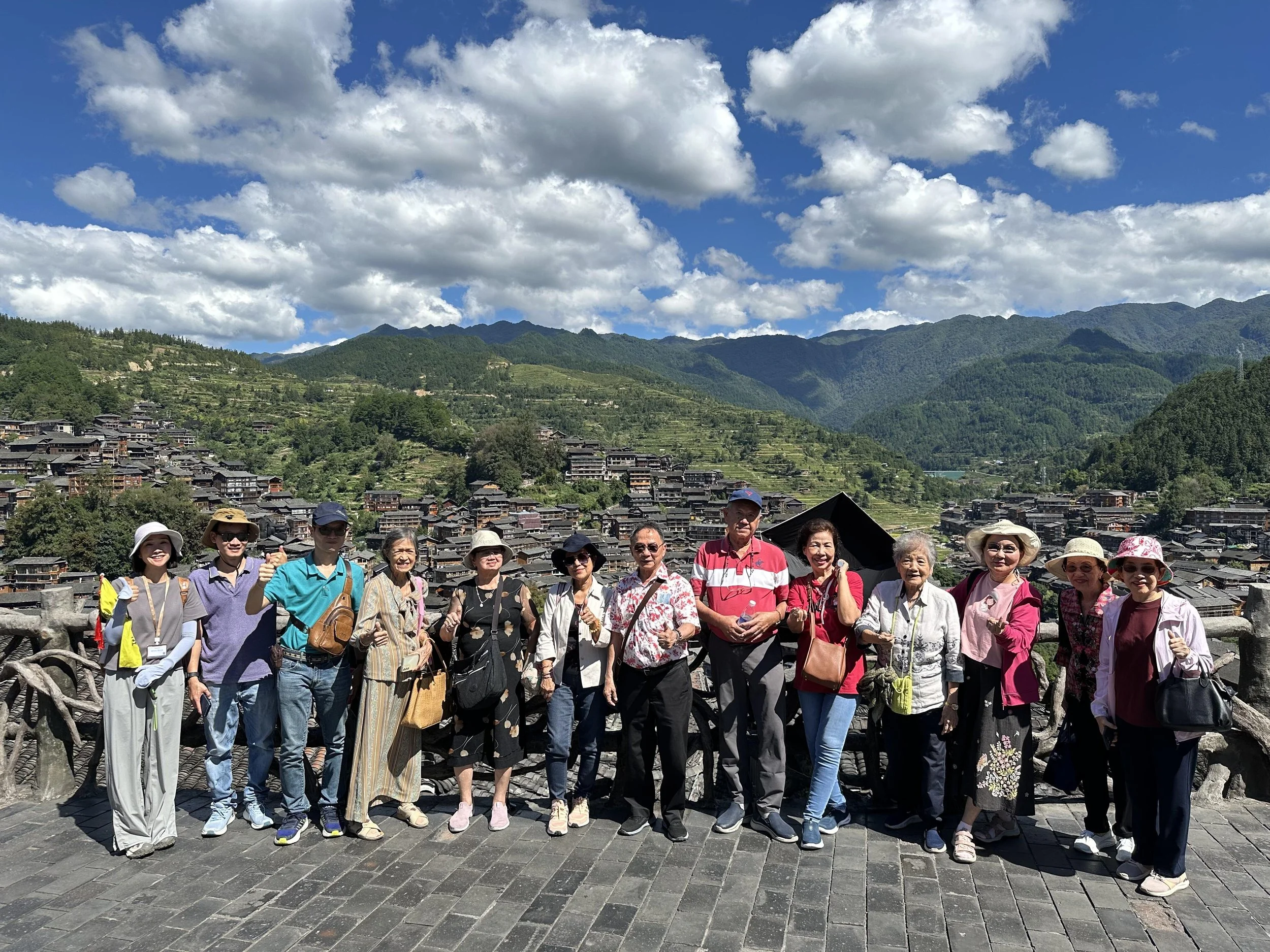 Retired professors from Maejo University in Chiang Mai, Thailand, posing with the stilt houses of Xijiang Miao Village in Guizhou during a cultural tour