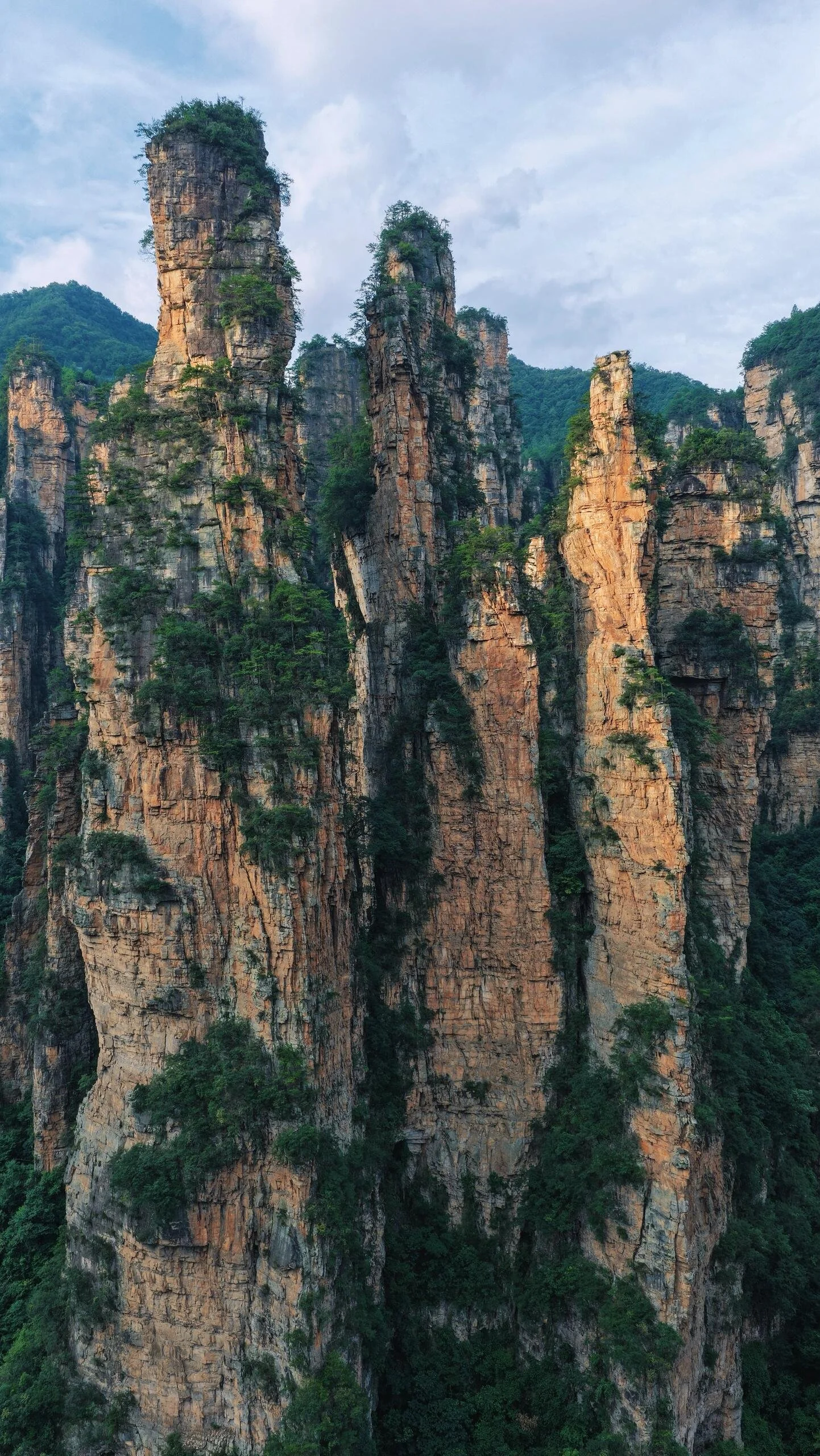 Tall, rugged sandstone pillars covered with sparse green vegetation in a mountainous landscape under a cloudy sky.