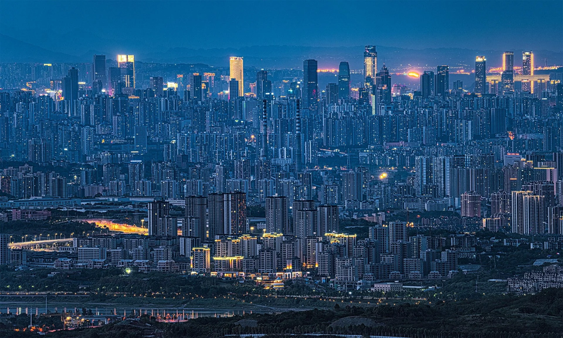 Nighttime cityscape of a dense urban area with numerous high-rise buildings illuminated, mountains in the background, and a river or body of water with reflections at the bottom.
