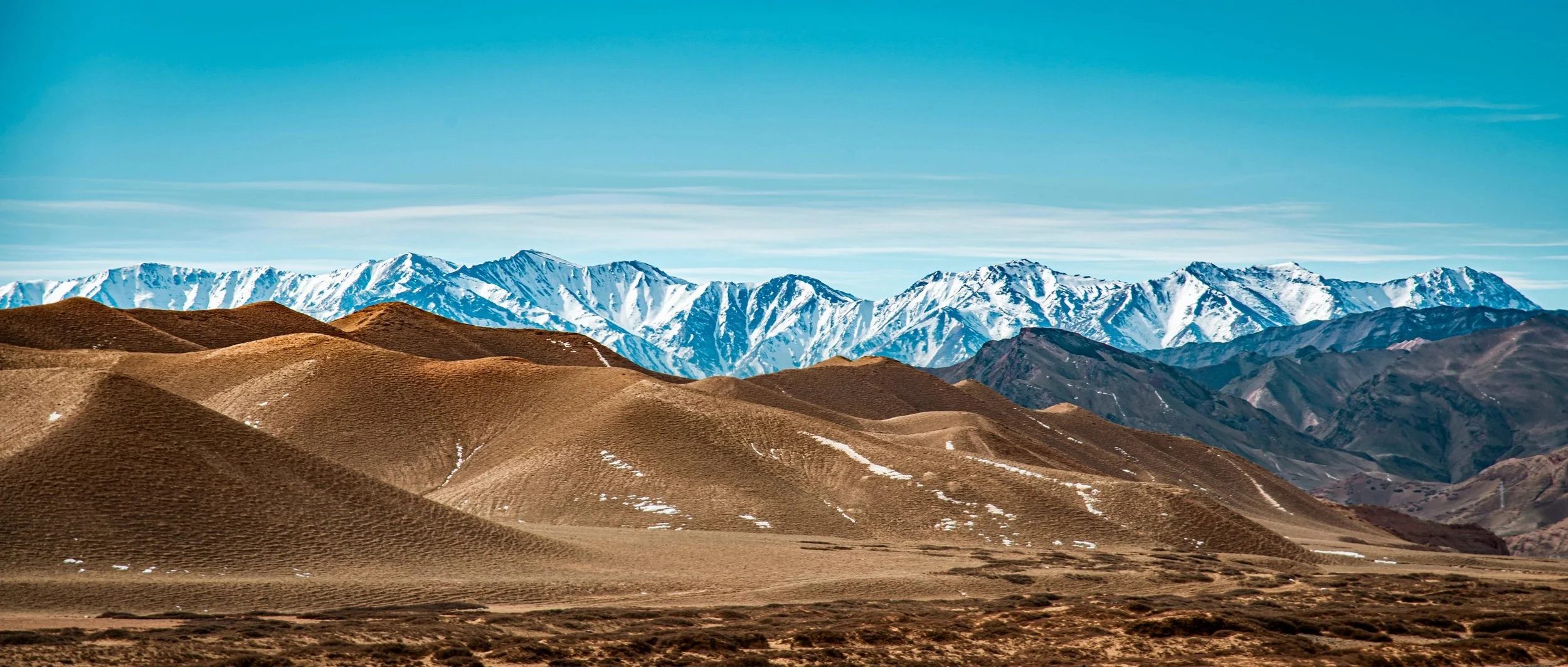 Scenic view of brown rolling hills in the foreground with snow-capped mountains in the background under a clear blue sky.