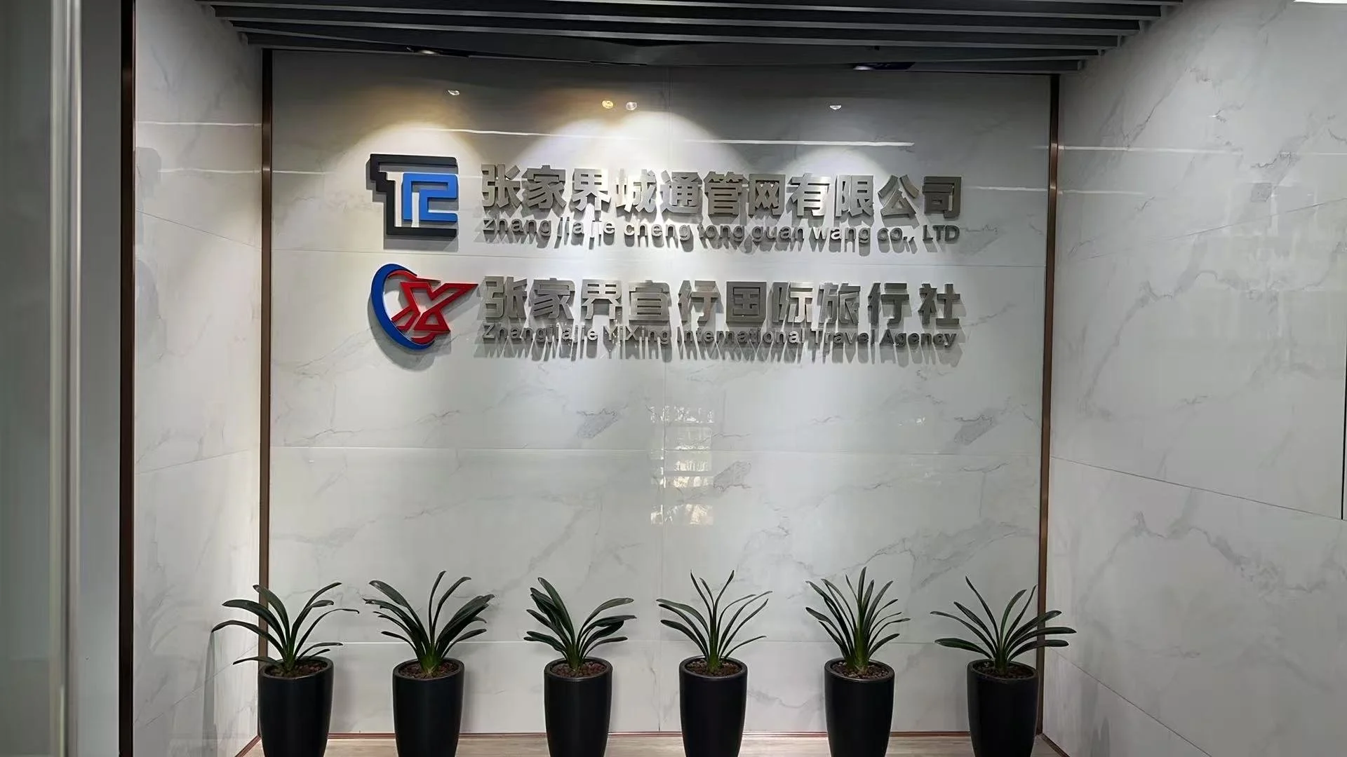 Company sign on marble wall with potted plants in front. The sign has Chinese characters and English translation for Zhang jia lie cheng tong guan wang CO., LTD and Zhang jia lie international travel agency.