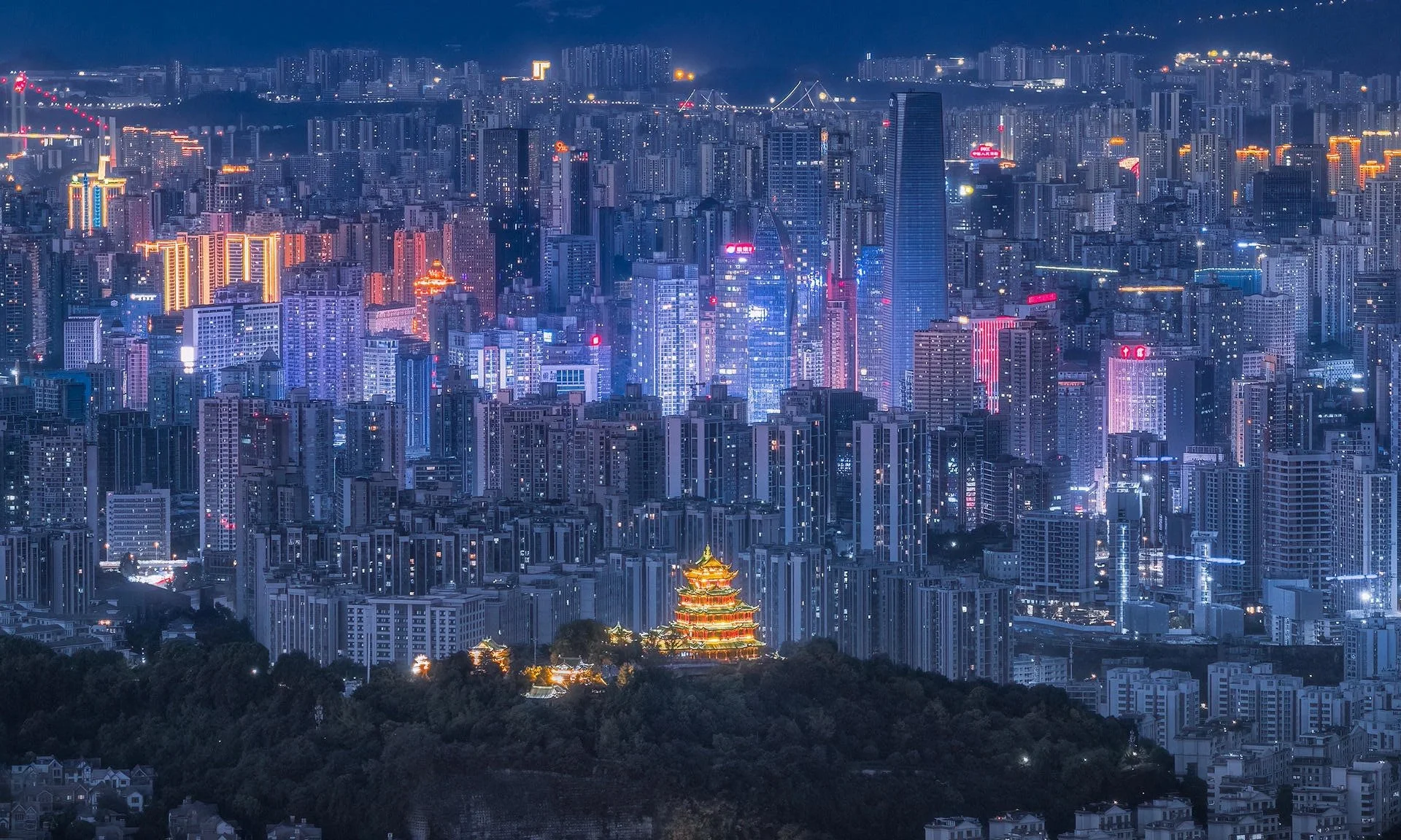 Nighttime cityscape with tall, illuminated skyscrapers, a brightly lit traditional Chinese pagoda in the foreground, and mountains in the background.