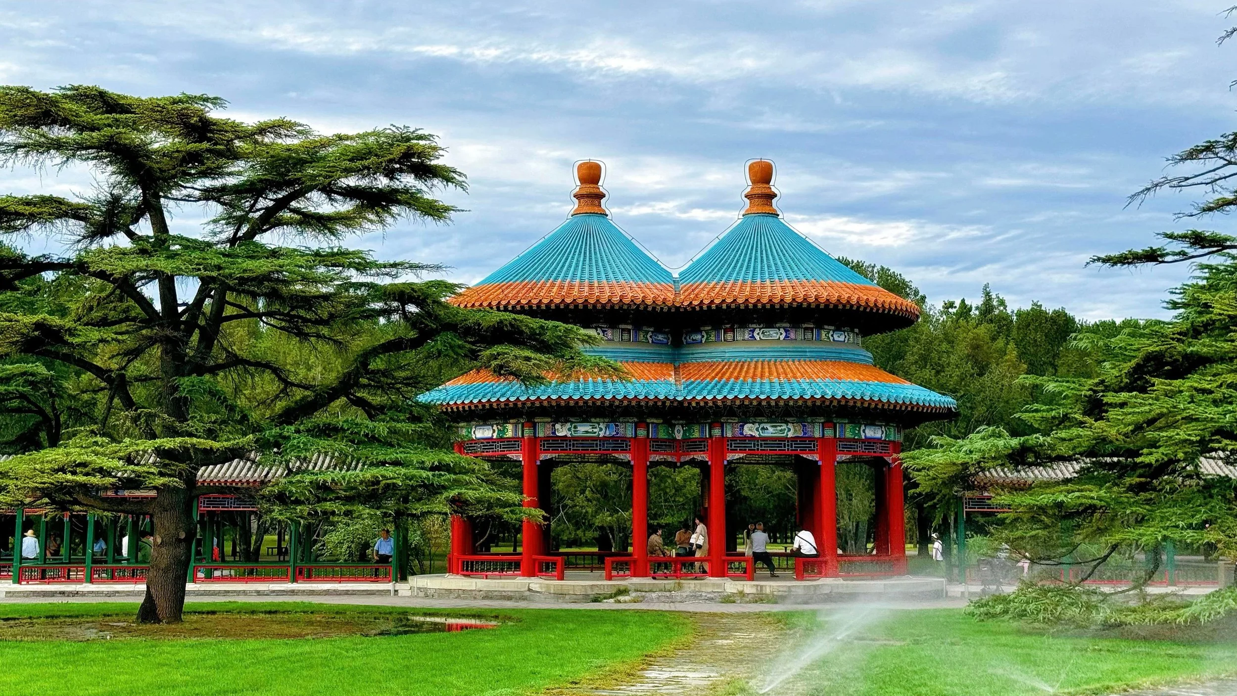 Photo of a traditional Chinese pavilion with a blue roof and red columns, surrounded by green trees and a park with people walking and sitting.