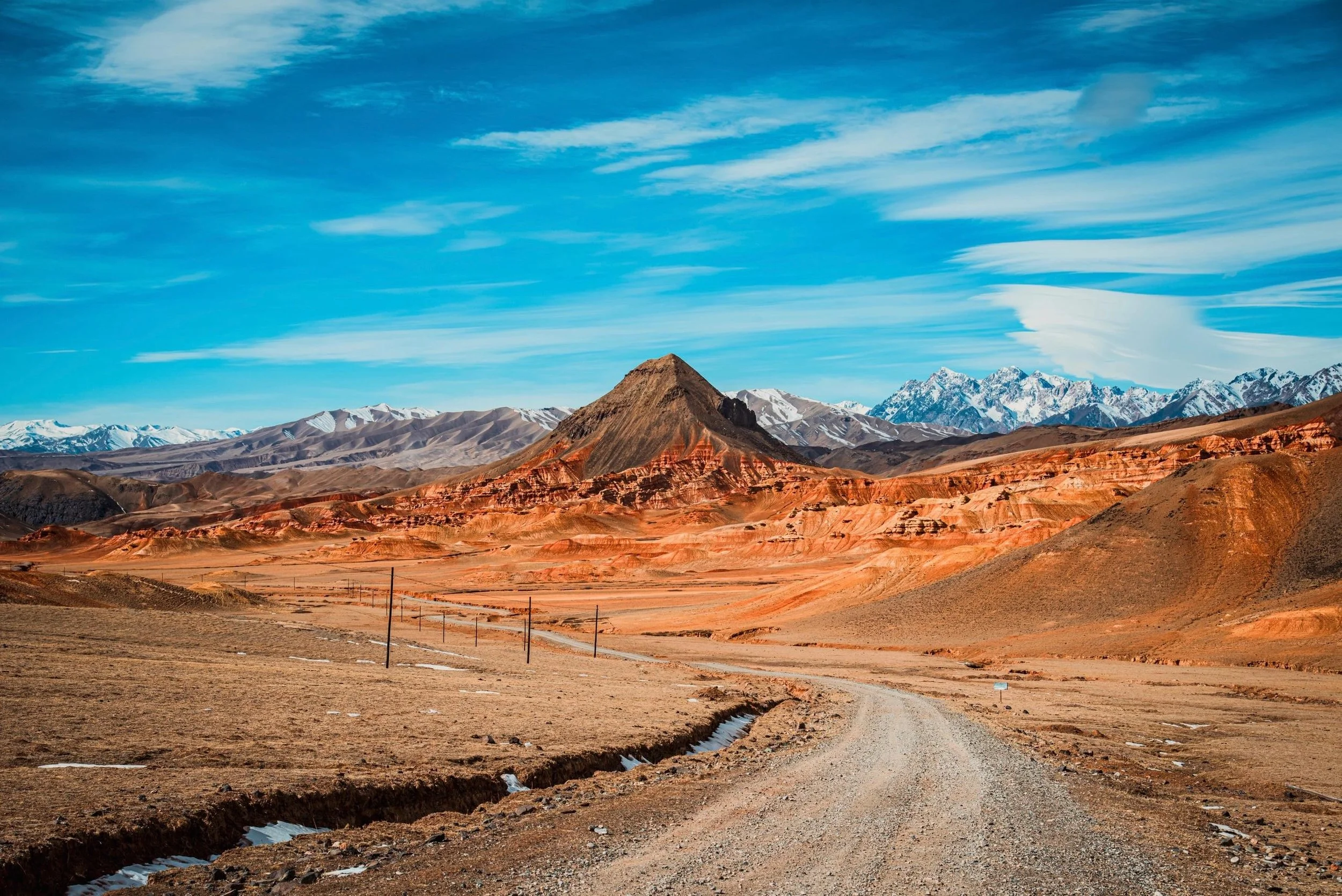 Desert landscape with colorful layered hills, mountain range with snow-capped peaks, and blue sky with wispy clouds.