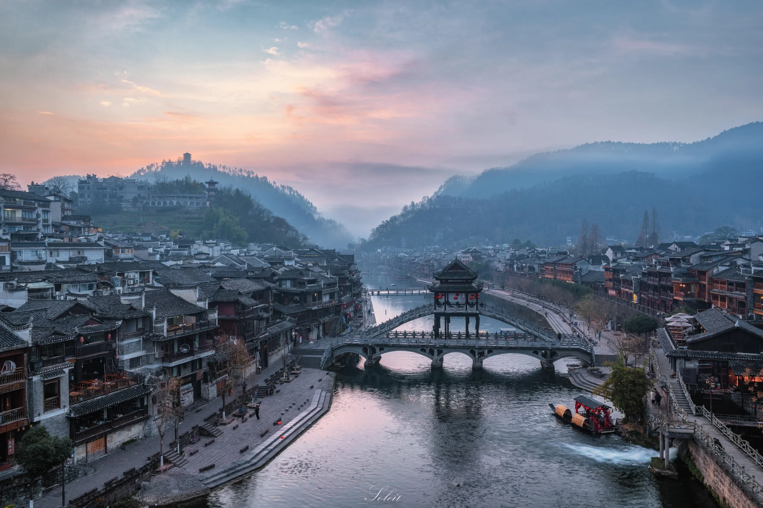A scenic view of a traditional Chinese water town with old buildings lining a river, a stone bridge crossing the water, mist-covered mountains in the background, and a colorful sky at dawn or dusk.