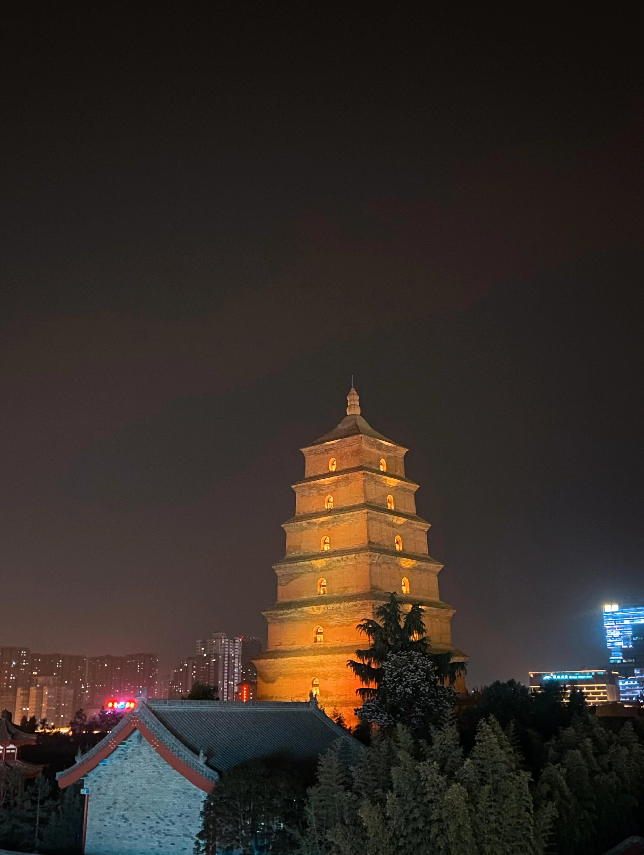 Night view of a traditional Chinese pagoda illuminated with warm yellow lights, surrounded by trees and modern buildings with bright city lights in the background.