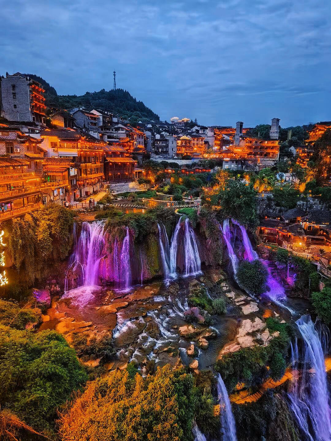 A cityscape at dusk with illuminated buildings on a hillside and waterfall with purple and white lights in the foreground.