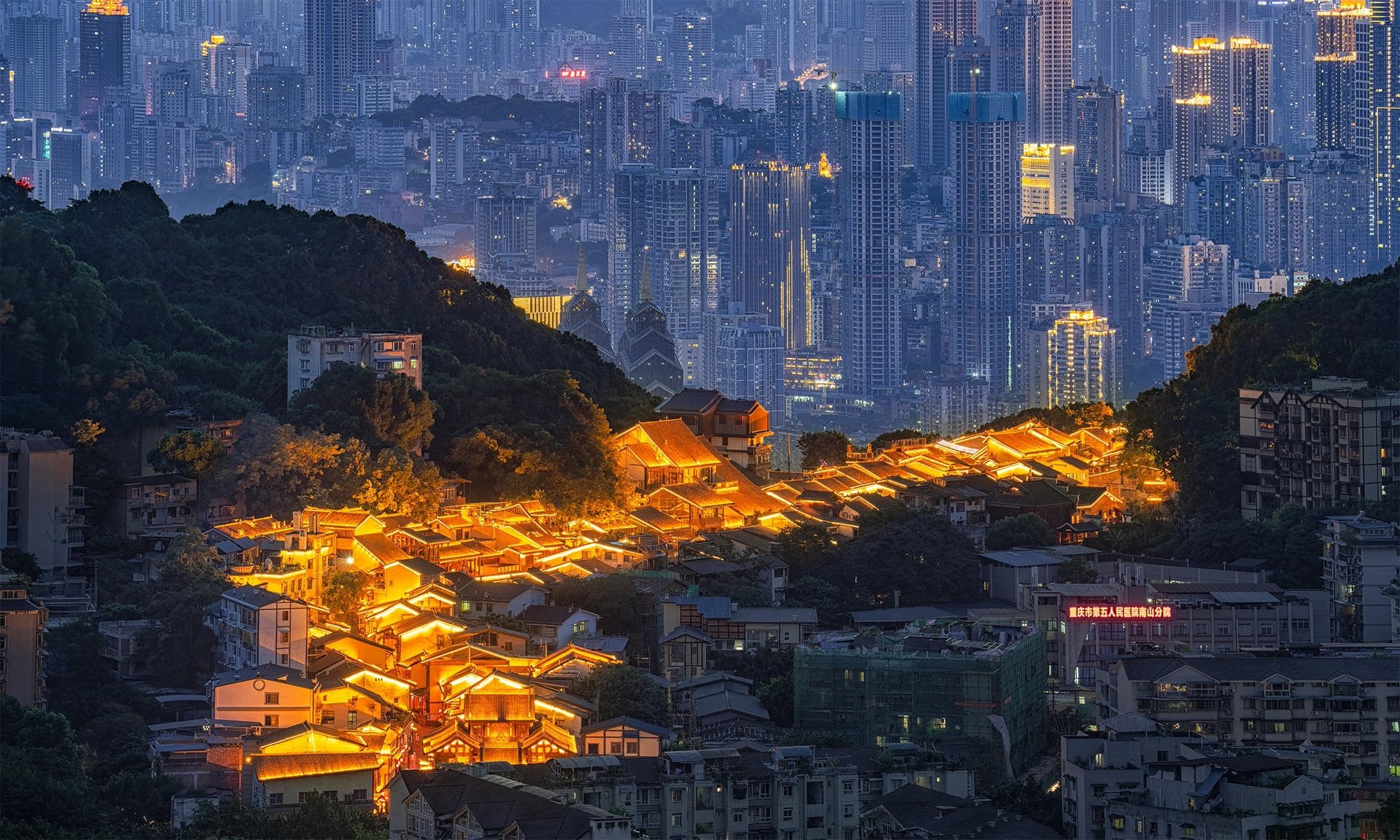 Nighttime cityscape with illuminated traditional-style houses at the forefront and modern skyscrapers in the background.