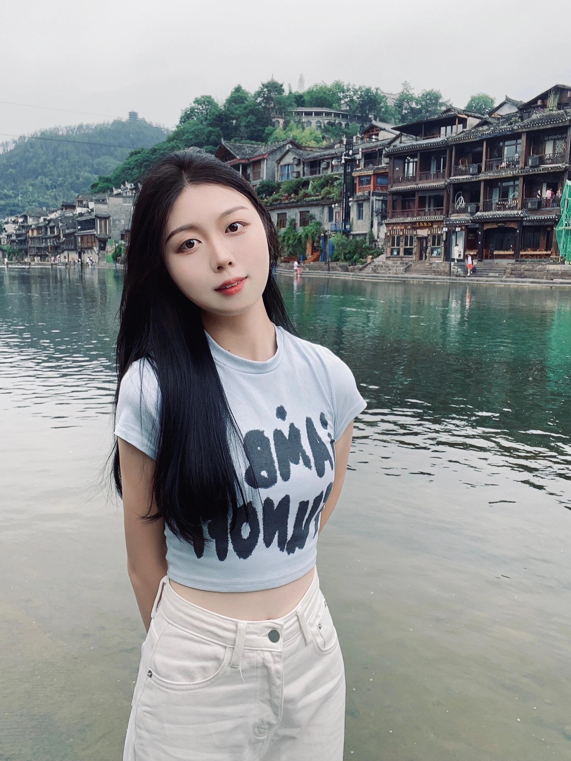 A young woman with long black hair standing by a river with traditional wooden houses on the hillside in the background.