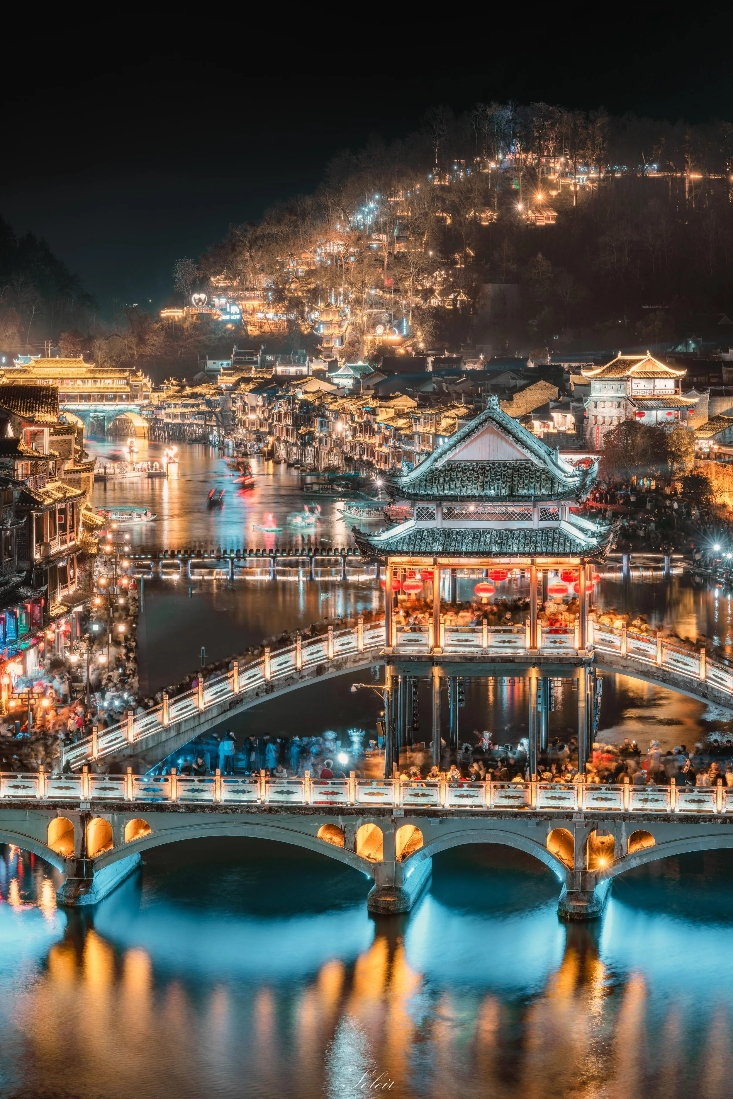 Night scene of a lit-up traditional Chinese bridge over a river, with a pagoda-style pavilion above the water, surrounded by illuminated buildings and a hillside with numerous houses and lights in the background.