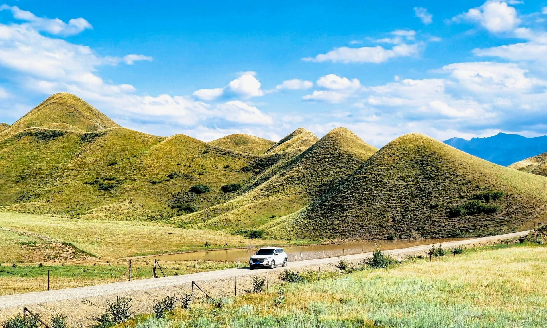 Green rolling hills under a partly cloudy blue sky with a white car driving on a dirt road in the foreground.