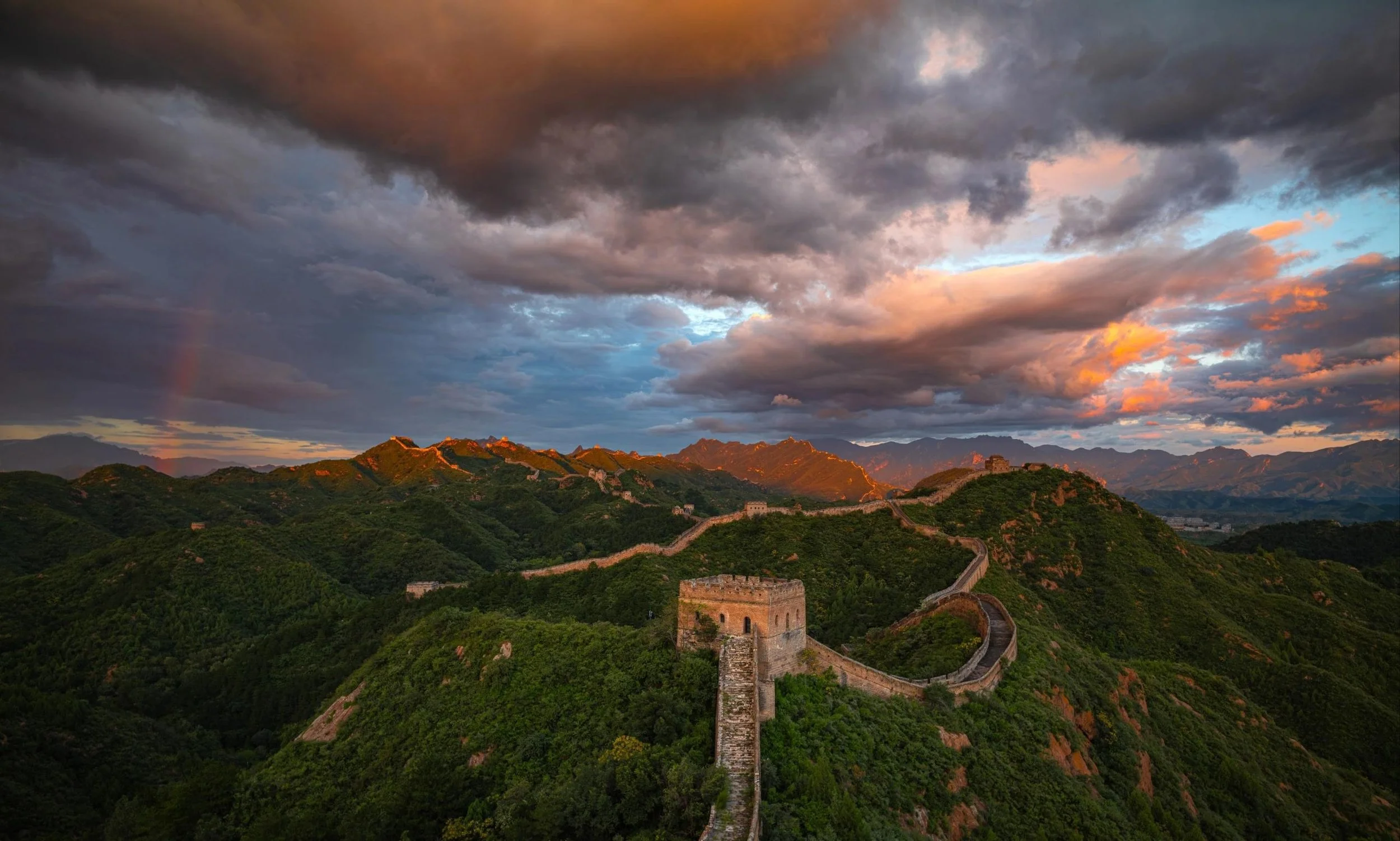 Sunset over the Great Wall of China winding through green mountains under a dramatic sky with dark clouds and hints of orange and blue.