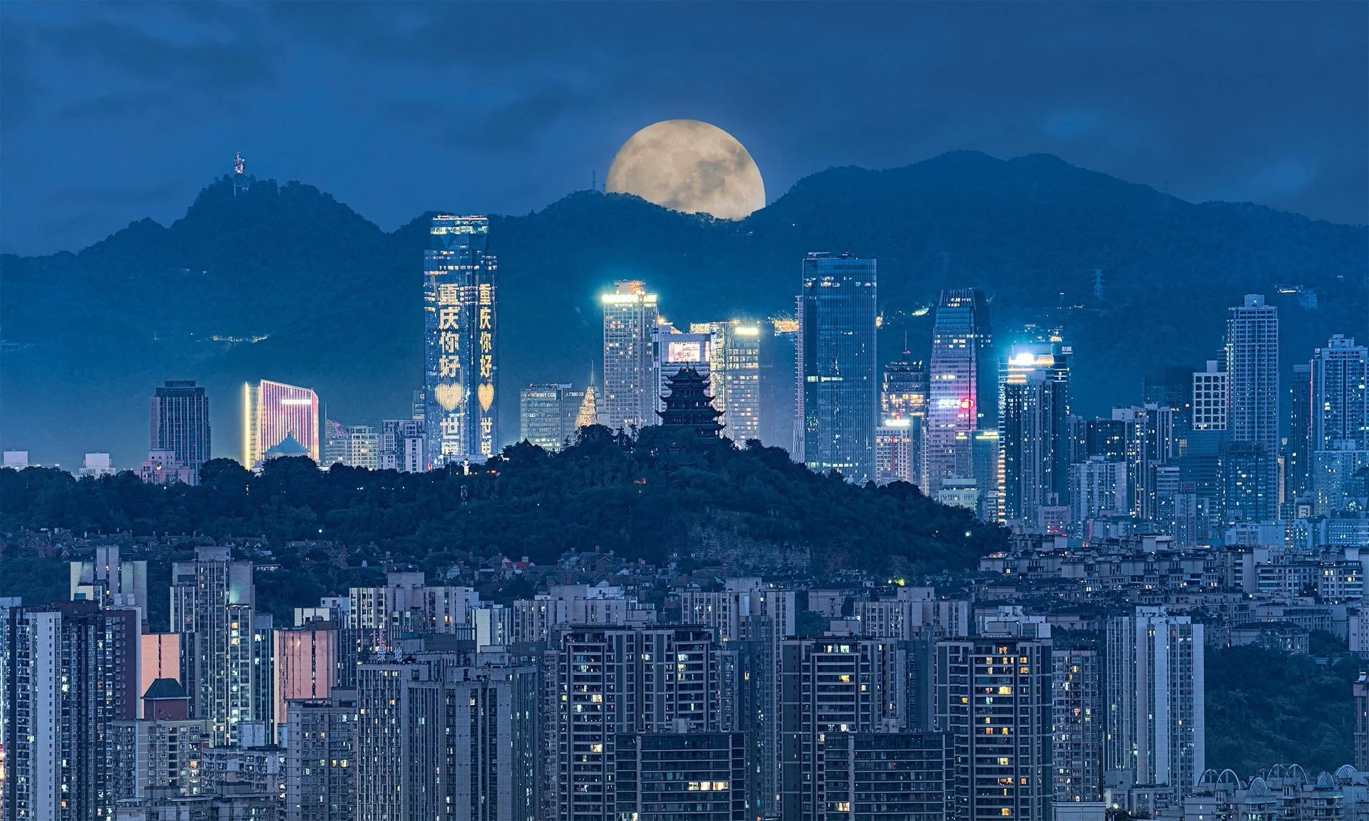 Nighttime city skyline with illuminated skyscrapers, a mountain range in the background, and a large full moon rising over the mountains.