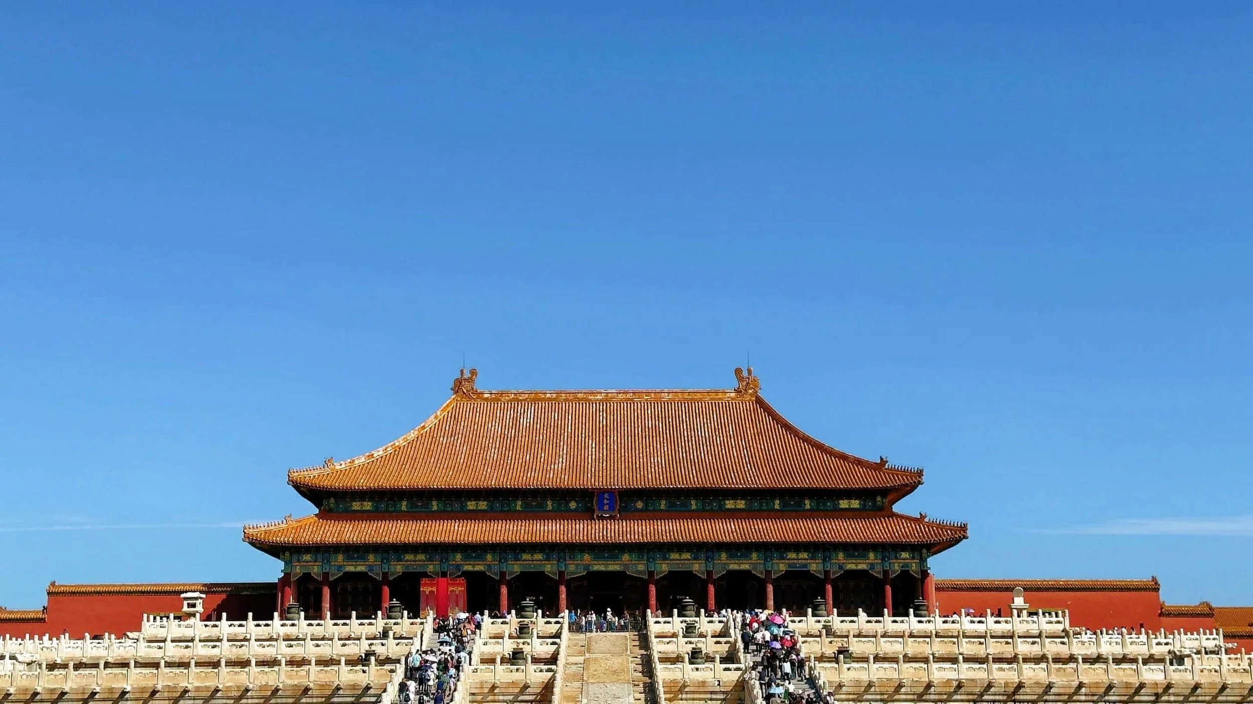 Panoramic view of a historic Chinese palace complex with traditional orange rooftops, green trees, and modern buildings in the background during sunset with orange and yellow sky