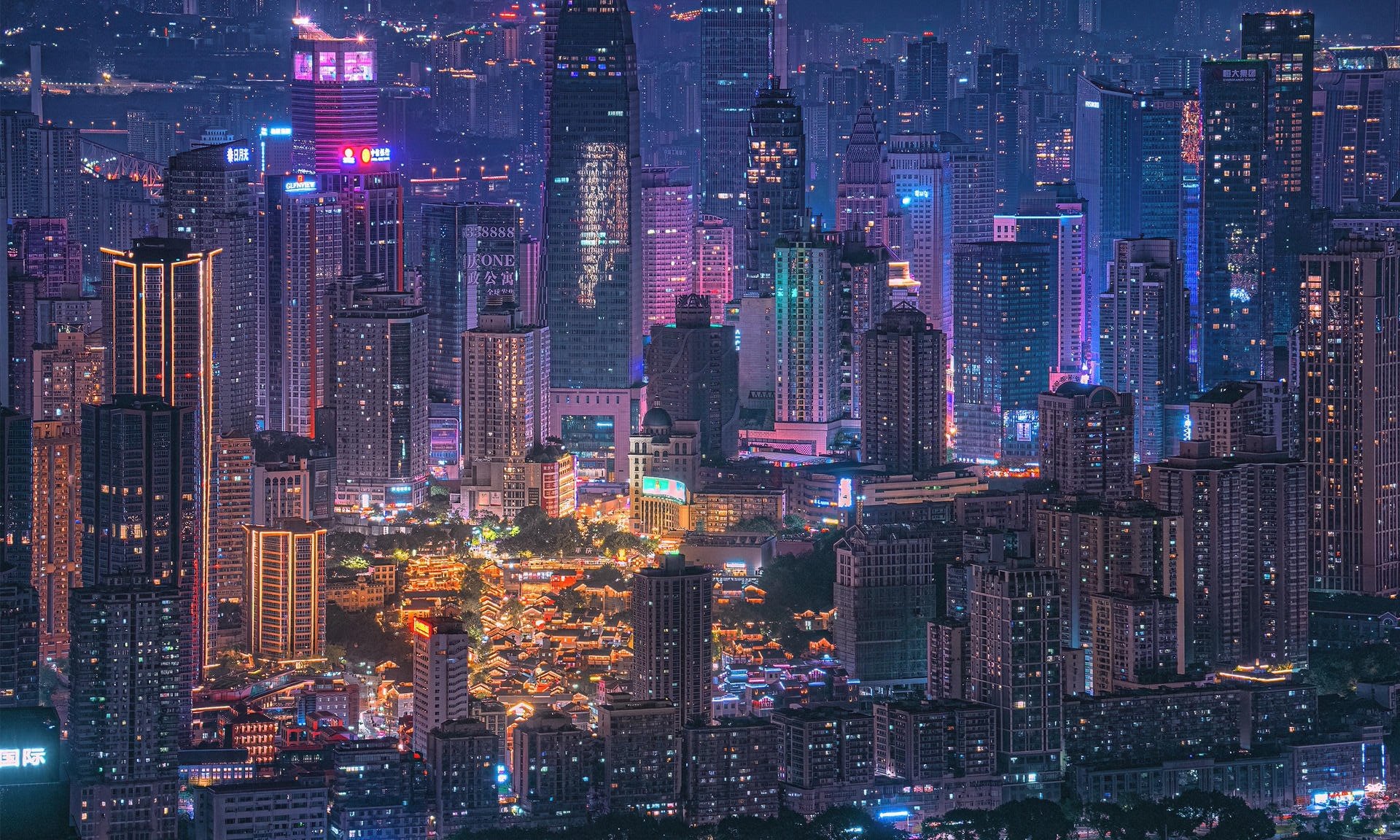 Nighttime view of a city skyline filled with illuminated tall skyscrapers and buildings, with colorful neon lights and signs, and a busy street scene at the bottom.