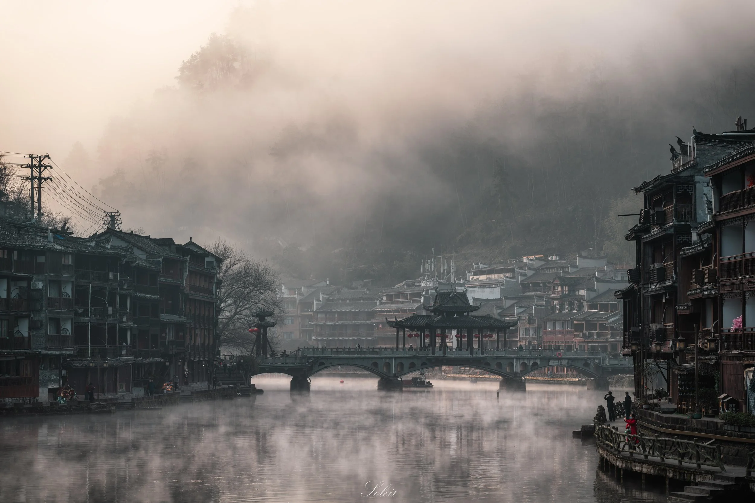 A foggy riverside scene with traditional Asian-style buildings, a stone bridge, and people enjoying the view on the riverbank.