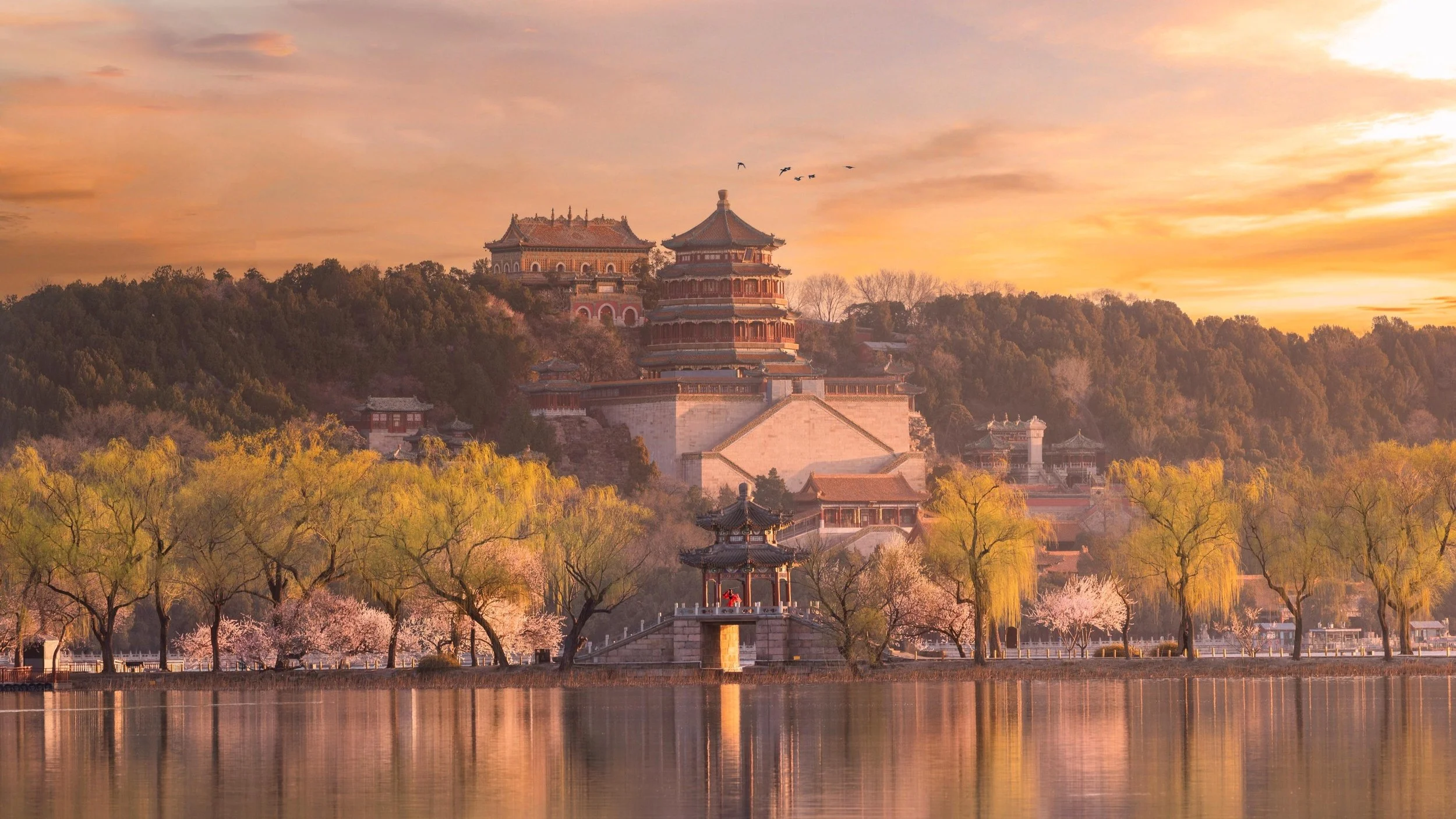A traditional East Asian temple complex on a hill, with pagoda-style buildings surrounded by trees, reflecting on a lake at sunset.