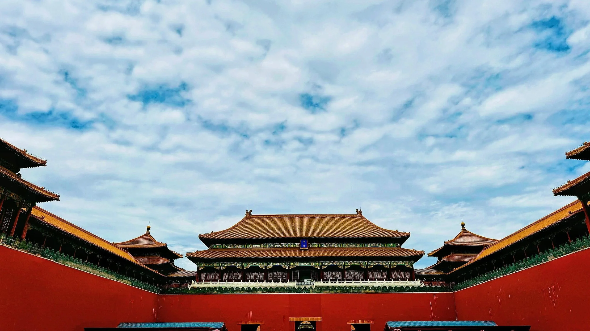 View of traditional Chinese palace buildings with orange-tiled roofs, surrounded by a red wall, under a blue sky with scattered clouds.