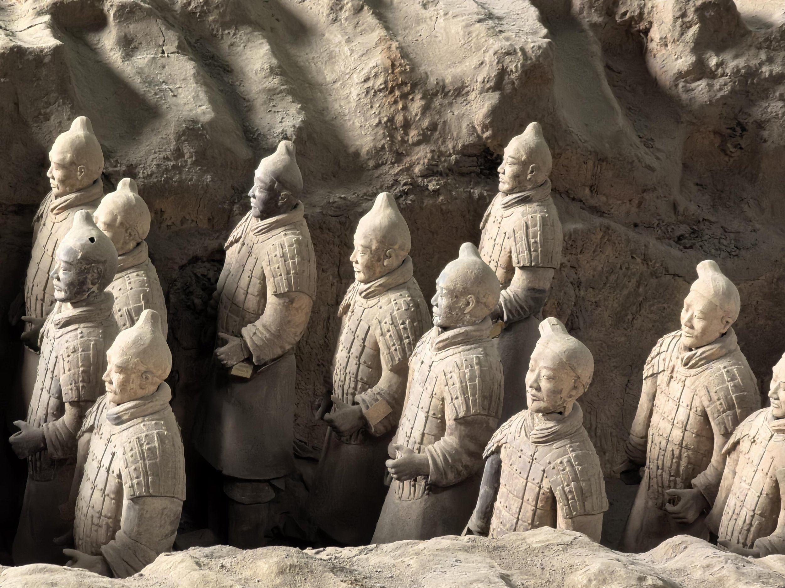 Close-up of the Terracotta Army statues depicting soldiers in battle formation been carved from clay or terracotta, with detailed facial features, armor, and hairstyles, set against a rocky background.