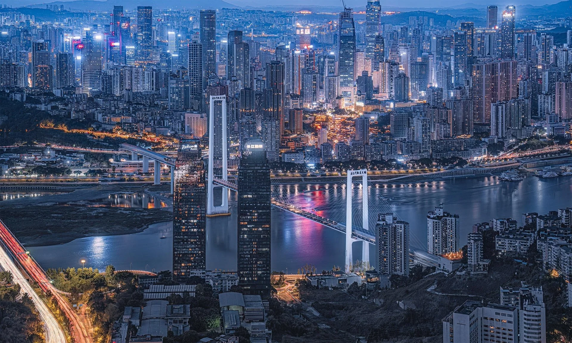A city skyline at night with numerous tall skyscrapers illuminated, a river running through the city, and a bridge crossing the river. The city is densely built with lights reflecting on the water.