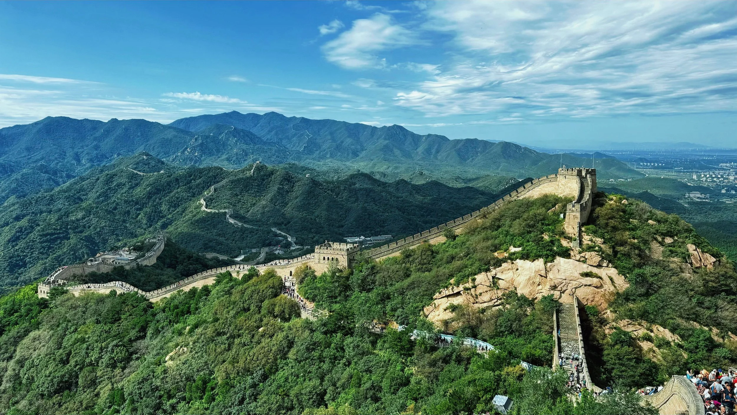Panoramic view of the Great Wall of China winding through lush green mountains under a blue sky with scattered clouds.