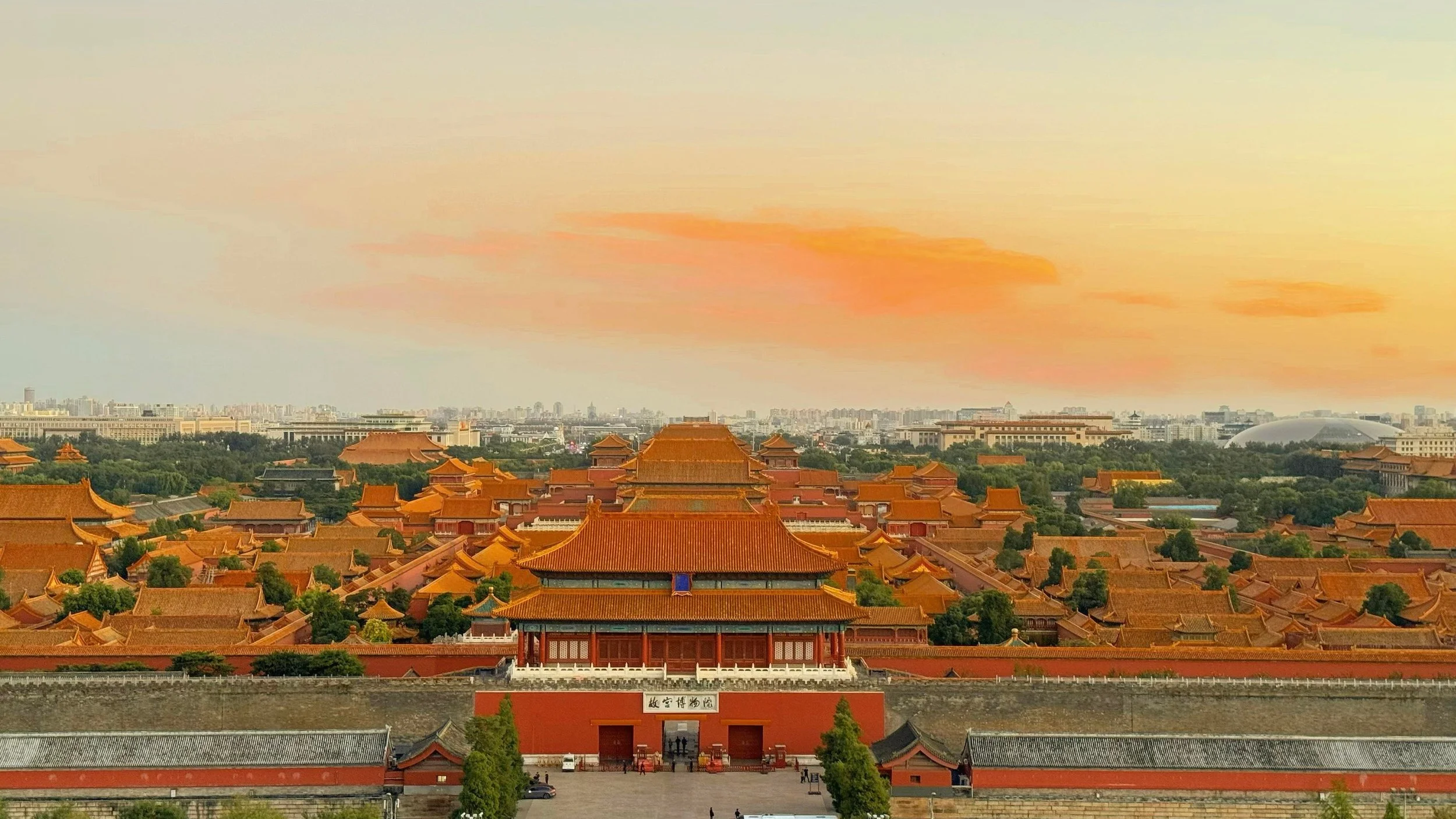 Panoramic view of a historic Chinese palace complex with traditional orange rooftops, green trees, and modern buildings in the background during sunset with orange and yellow sky