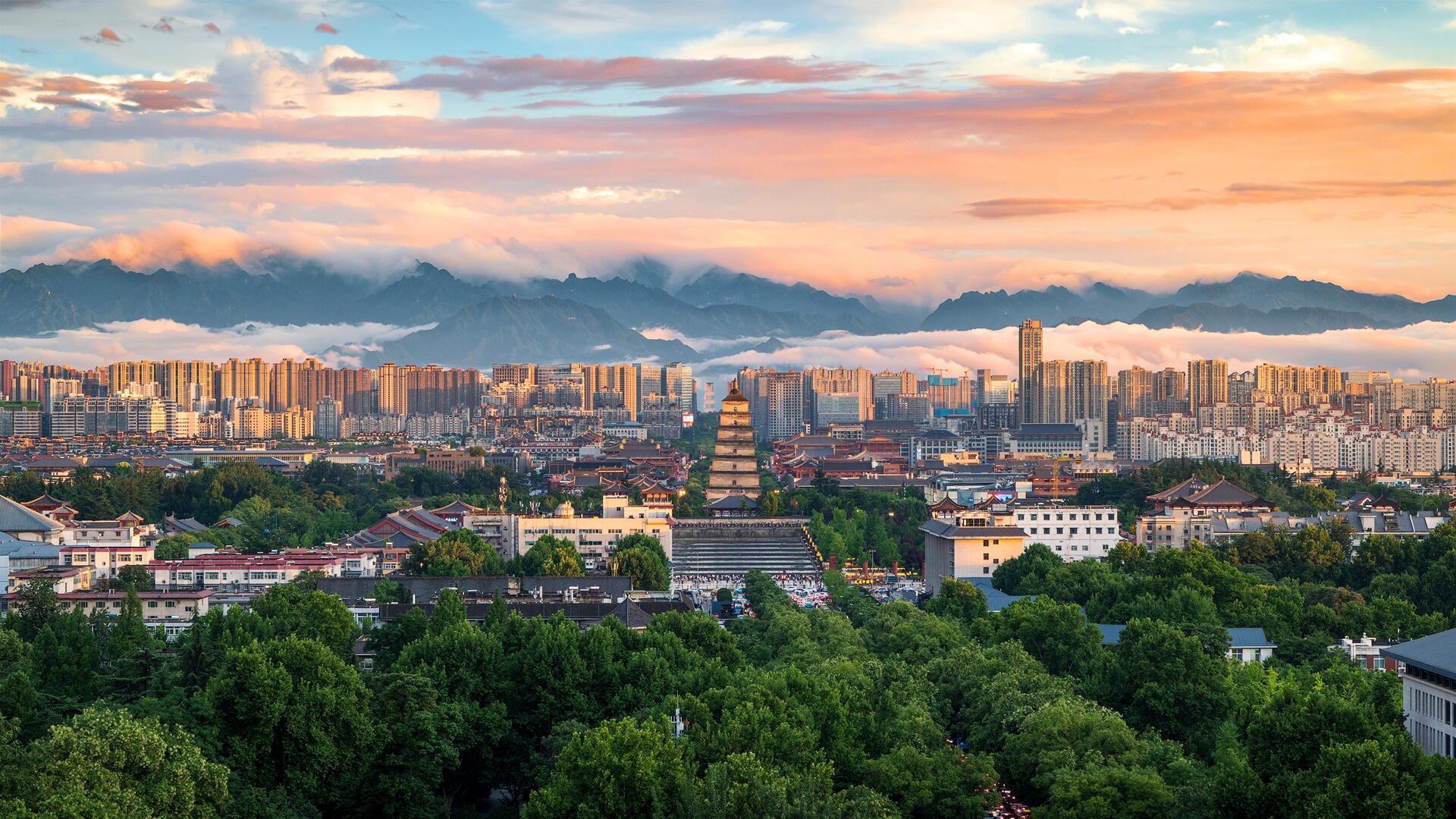 City skyline with high-rise buildings, traditional pagoda, lush green trees, and mountains in the background during sunset.