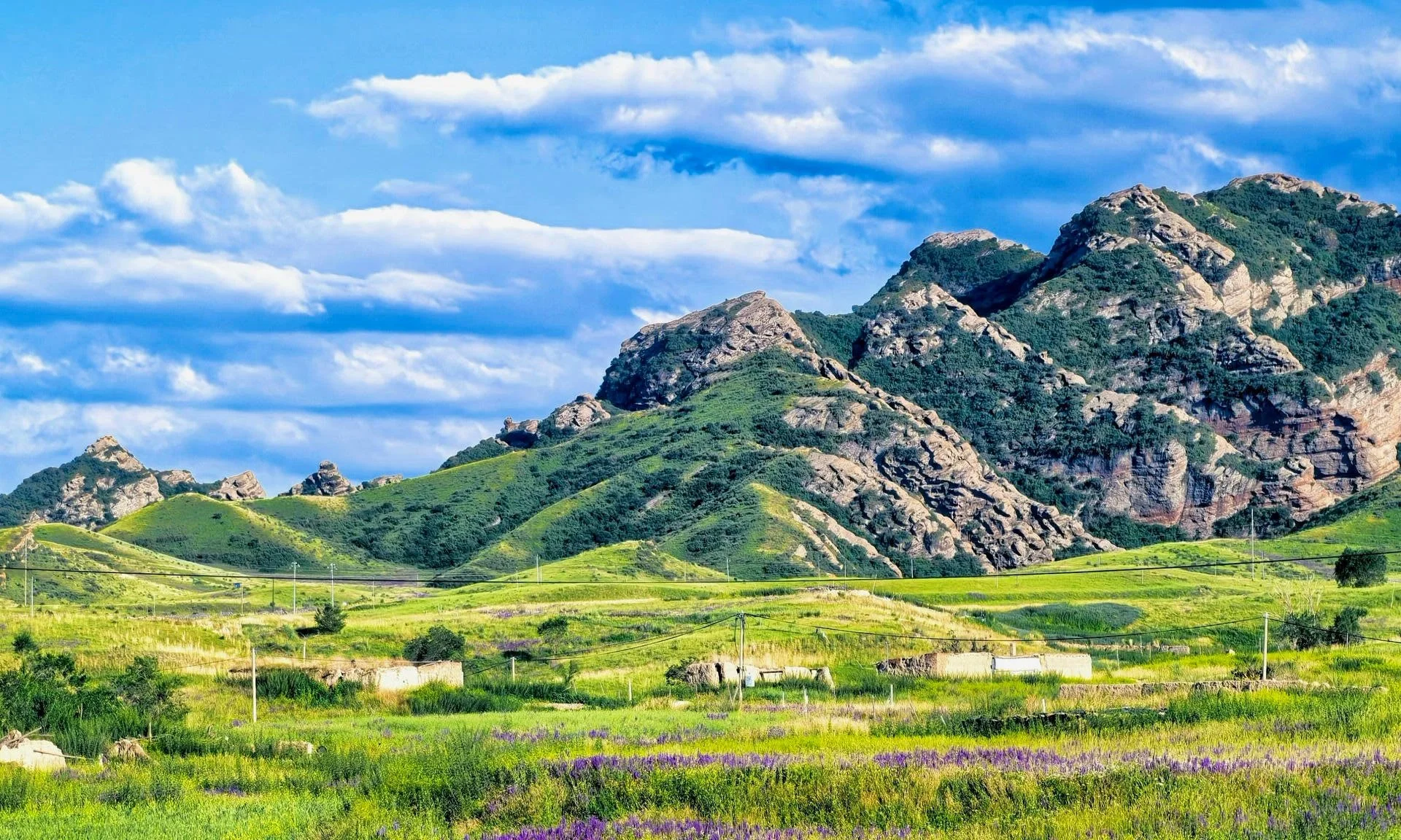 A scenic landscape of green hills and rocky mountains under a partly cloudy sky with blue and white clouds.