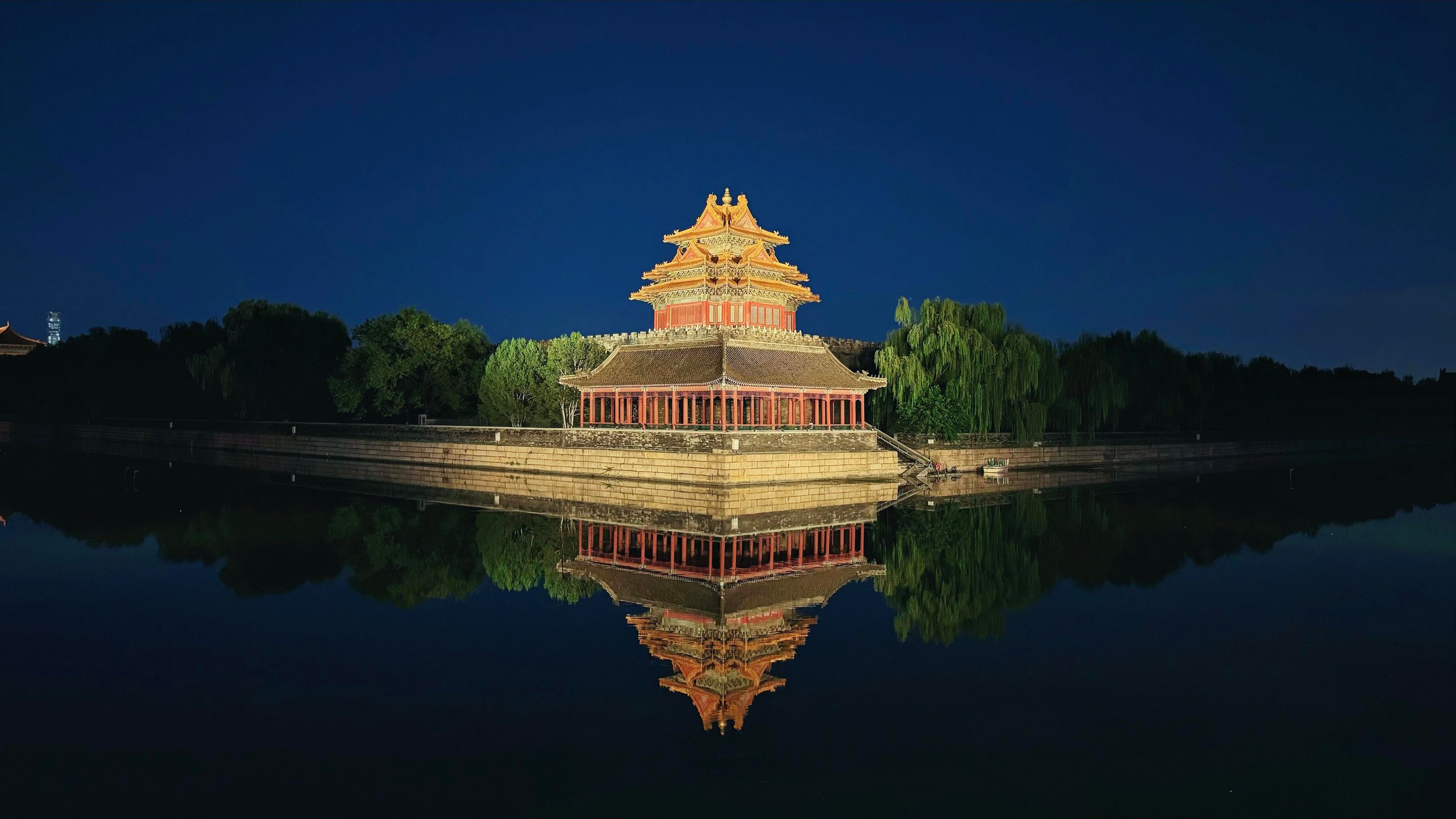 Nighttime view of a traditional Chinese pagoda with multiple tiers and ornate roof decorations, reflected in the water below, surrounded by trees and a clear dark sky.