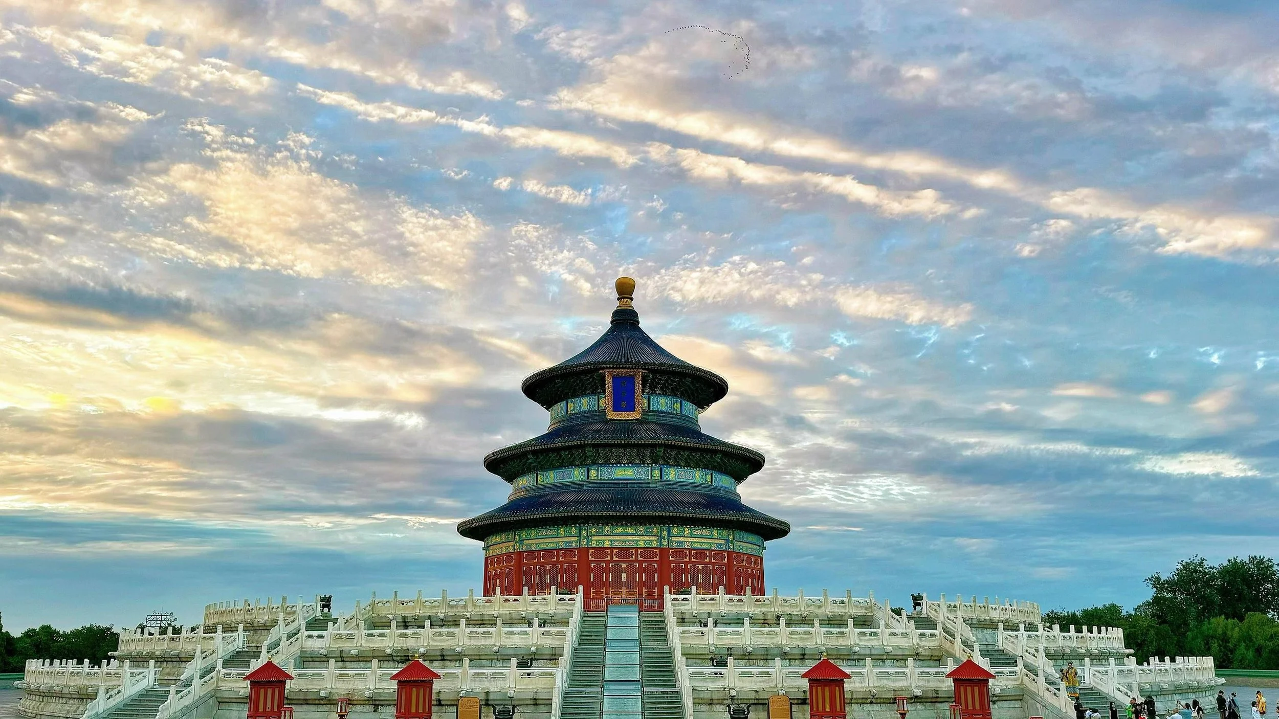 The Temple of Heaven in Beijing, China, with a multi-tiered circular roof, intricate decorative details, and surrounded by white stone railings and stairs, under a partly cloudy sky at sunset.