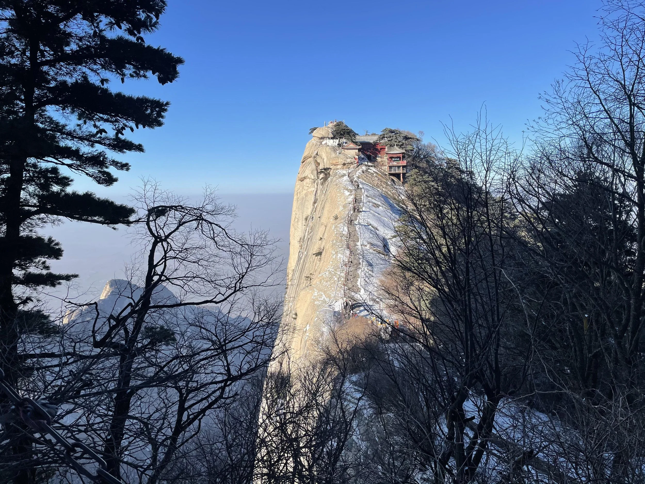 A mountain peak with temples and structures built on top, surrounded by trees and snow, under a clear blue sky.