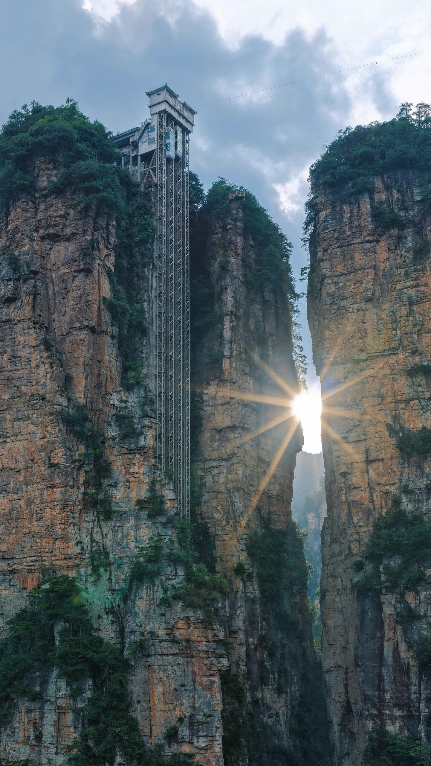 Elevator shaft on a tall mountain cliff with a cutout at the bottom, sunlight shining through the opening, and green trees on the rocky cliffs.