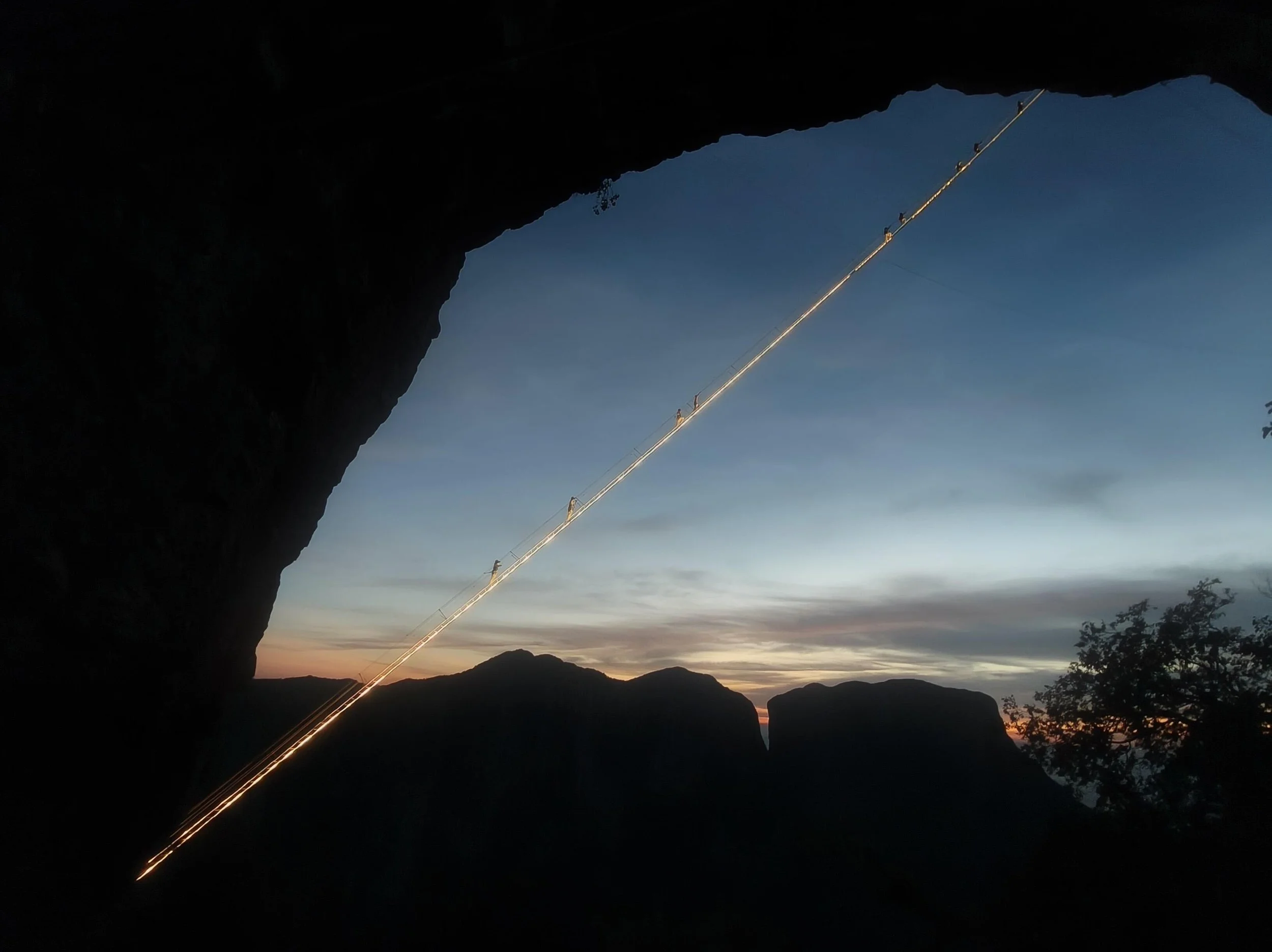 Panoramic view of the lit Sky Ladder via ferrata at dusk on Qixing Mountain Zhangjiajie, climbers appearing as small silhouettes along the glowing cable line above layered mountain ridges