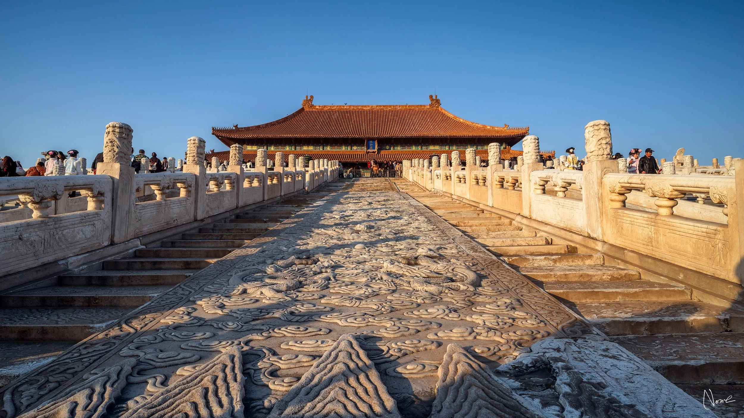 Traditional Chinese architecture building with a red tiled roof and ornate decorations, surrounded by a stone bridge with carved dragon designs, in a clear bright day with a blue sky, and people walking around.