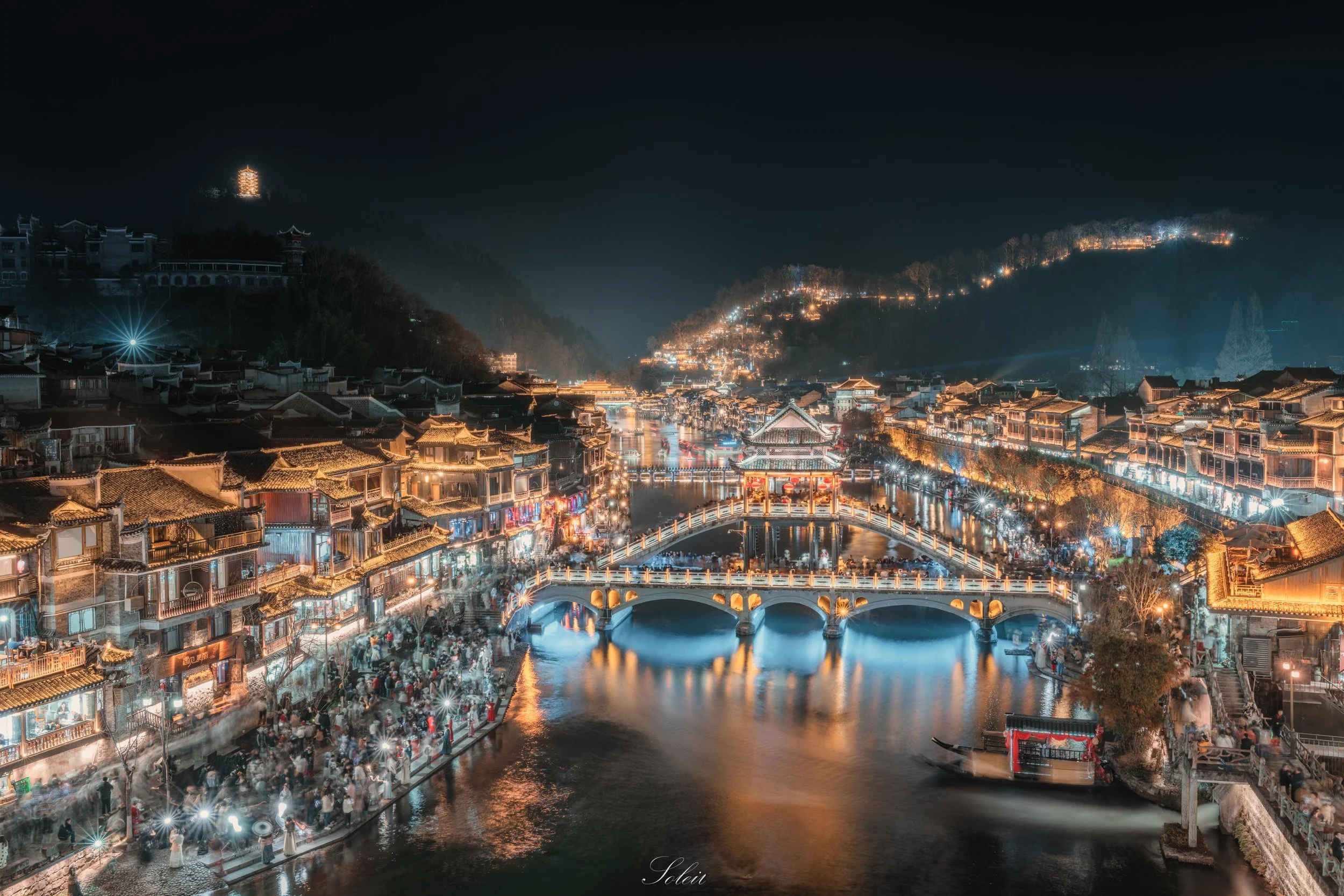 Night view of a traditional Chinese town with illuminated buildings, bridges, and a river, surrounded by hills with lit-up structures and a large pagoda in the distance.