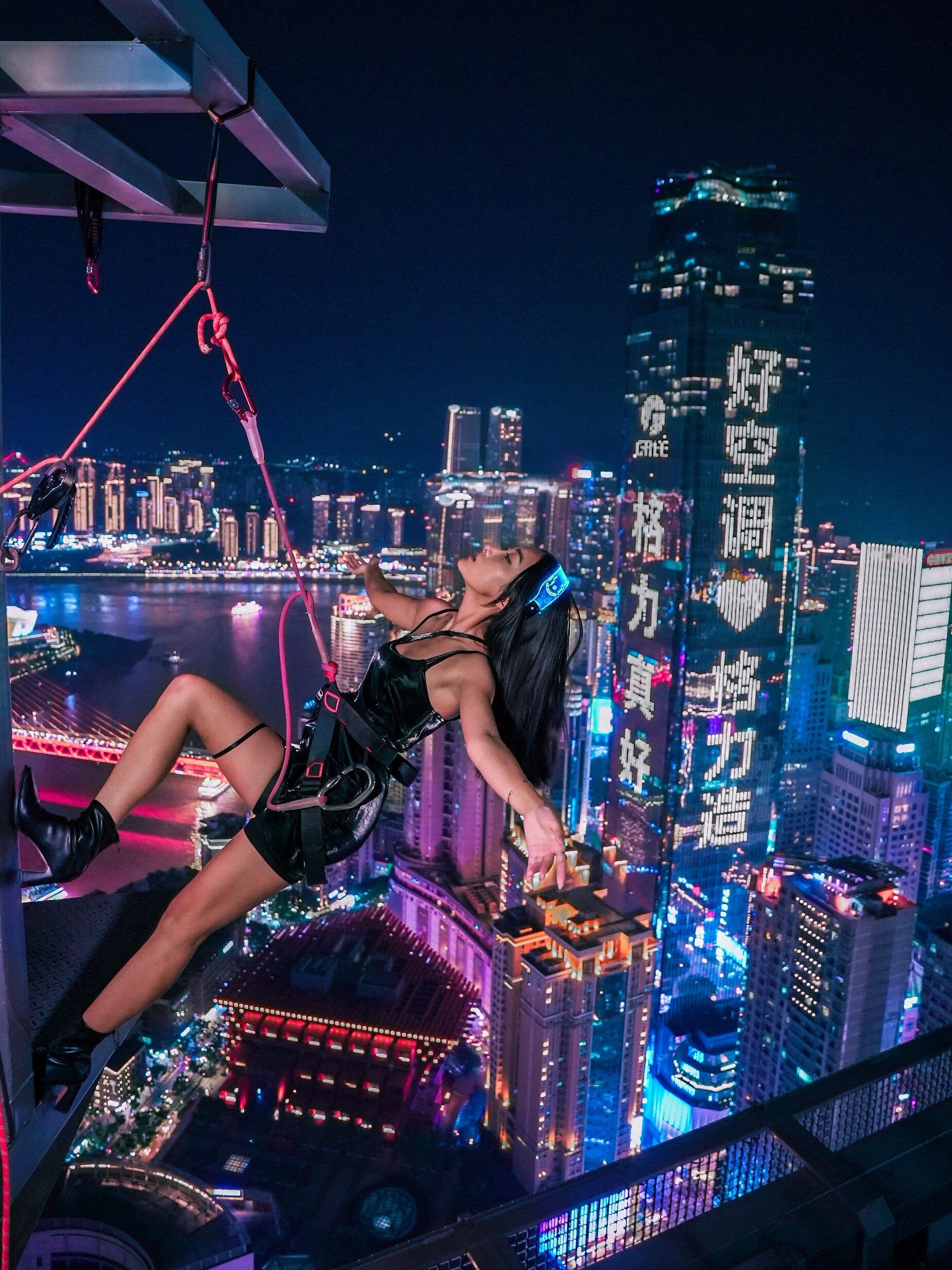 A woman sitting on a ledge overlooking a city skyline at night, with neon lights and tall buildings, some with illuminated signs, in the background. She is wearing a black outfit and headphones, with her head tilted back and arms outstretched.
