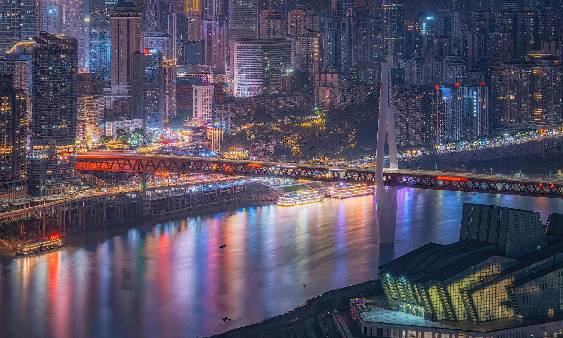 Nighttime cityscape of a dense modern metropolis with high-rise buildings, illuminated streets, and a river reflecting colorful lights, featuring a bridge with a unique cable-stayed design.