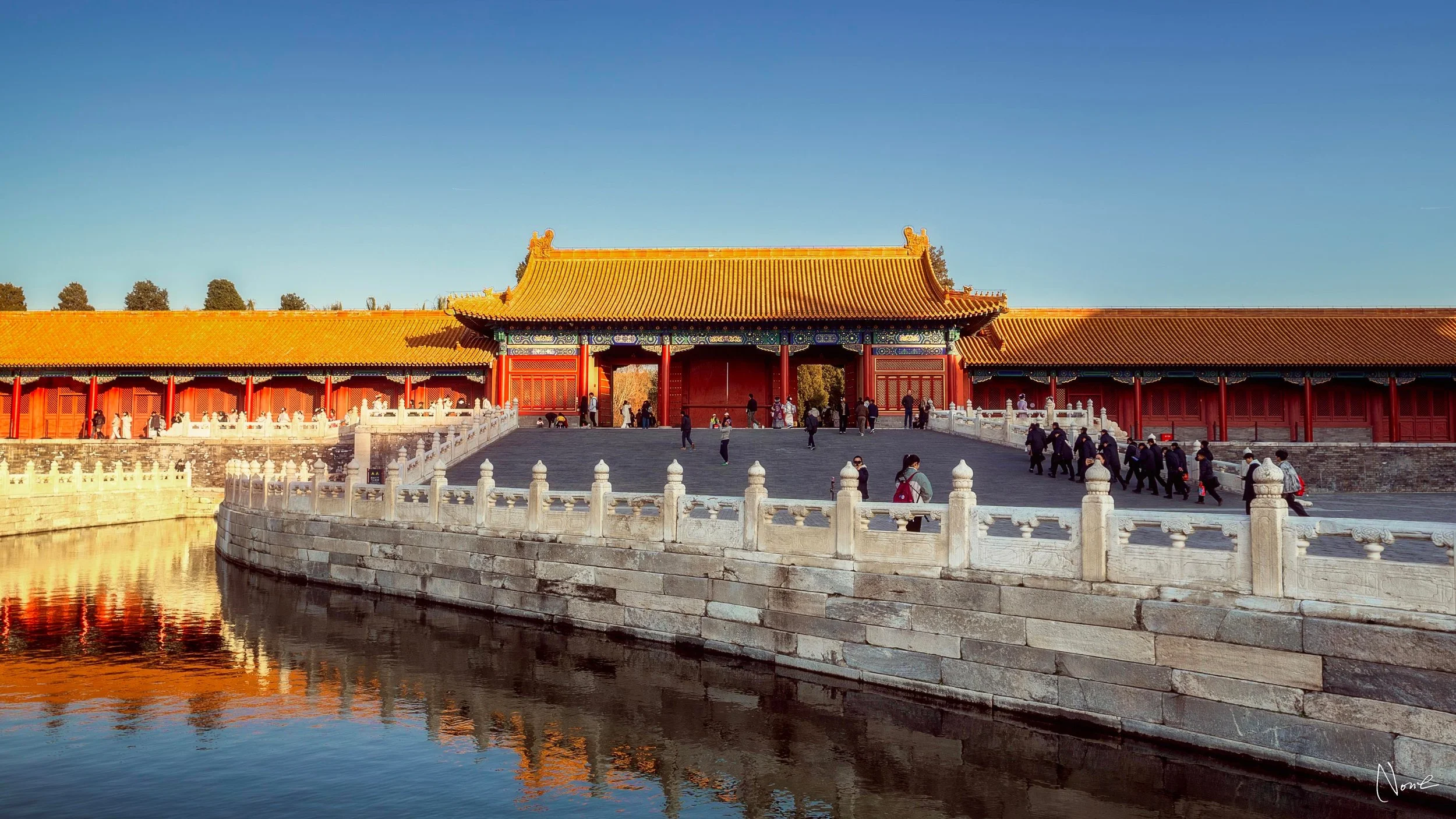 Traditional Chinese palace with red walls and yellow-tiled roof, visitors walking on stone pathway, water reflecting the structure, blue sky in the background.