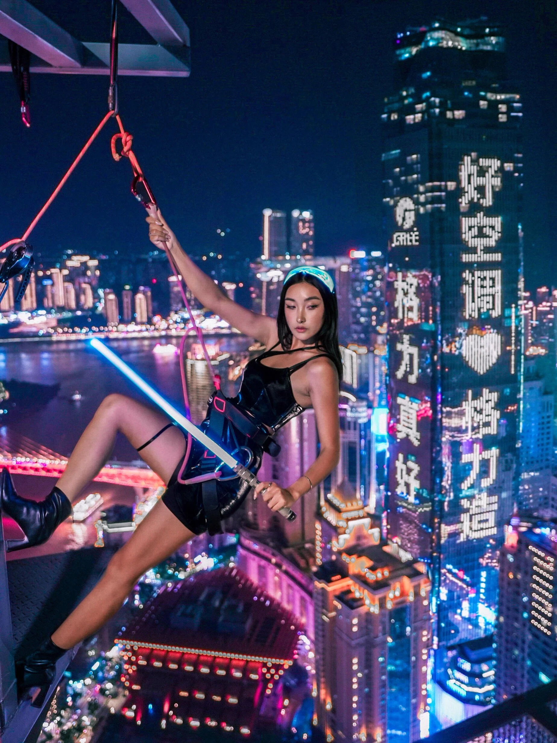 A woman in black attire, with a headband, hanging from a harness on a building rooftop at night with a city skyline and illuminated skyscrapers in the background.