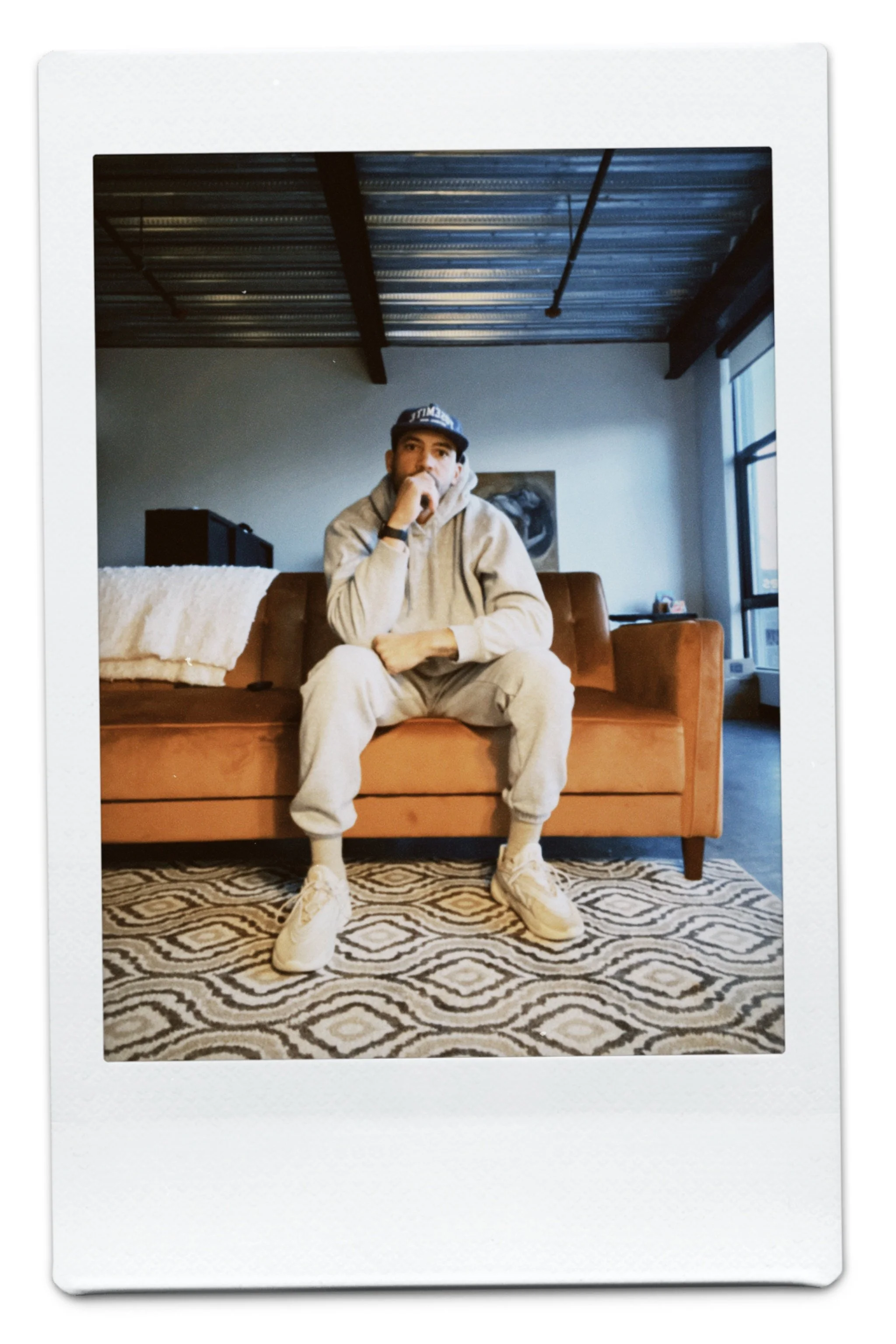 A young man is sitting on a brown couch in a modern living room with a patterned rug, wearing a gray hoodie, beige sweatpants, white sneakers, and a baseball cap. The room has a metal ceiling, a large window on the right, a piece of artwork on the wall, and a black piece of furniture in the background.