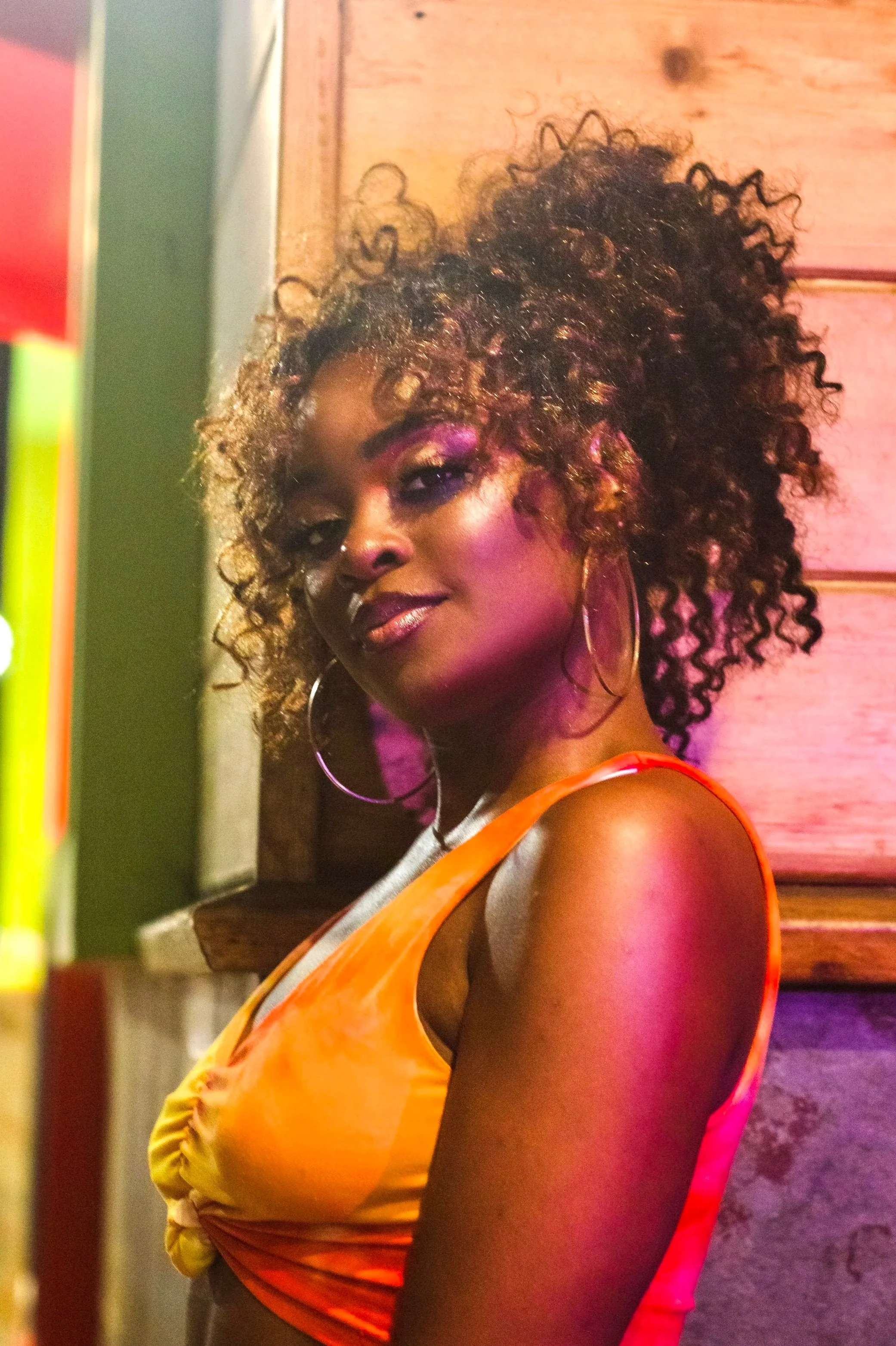 A woman with curly hair and large hoop earrings wearing an orange top, standing near wooden panels, lit with pink and purple lighting.