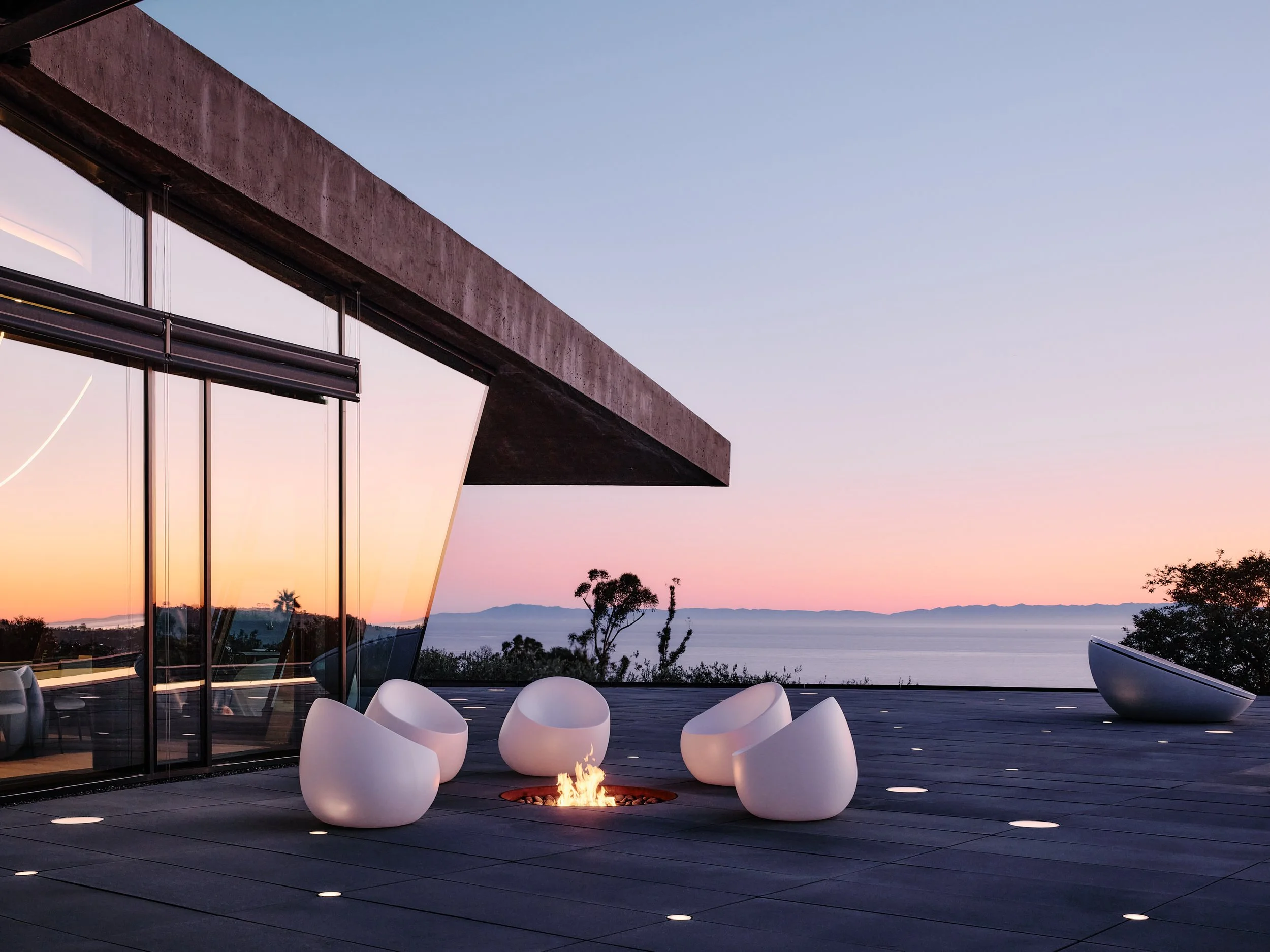 Modern house balcony overlooking water and mountains at sunset, with white round chairs and fire pit.
