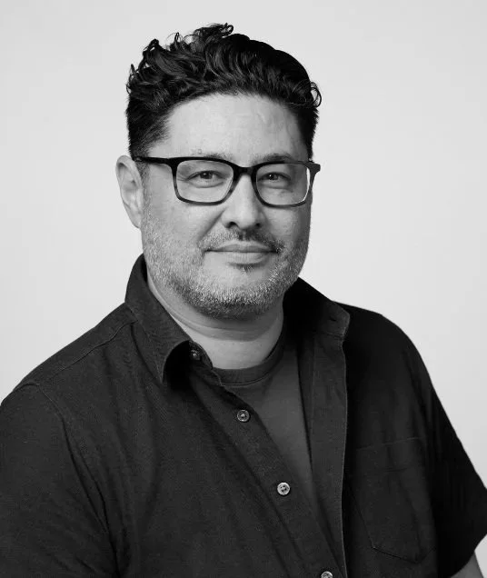 Black and white portrait of a man with glasses, short curly hair, and a beard, wearing a dark button-up shirt, smiling slightly against a plain background.