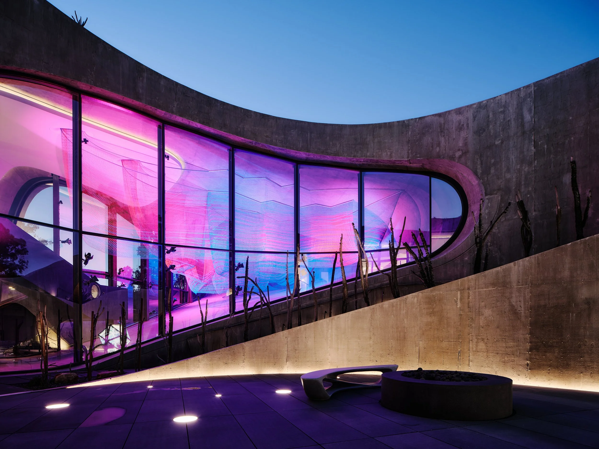 Modern building with large glass windows illuminated with pink and purple lights during early evening, featuring a curved architectural design and an outdoor seating area with benches and landscaped plants.