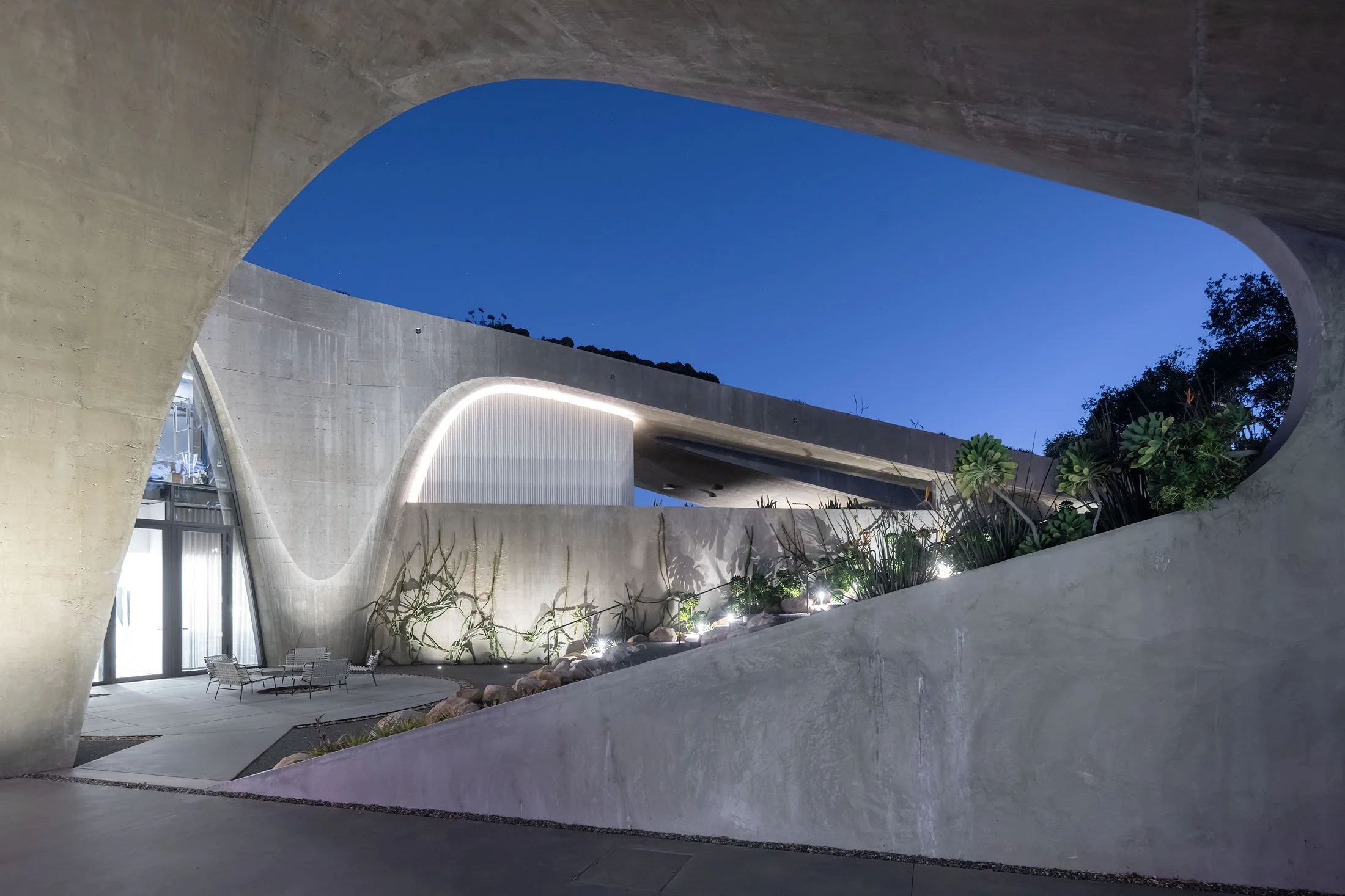 Modern architectural building with large concrete arches and curved walls. Nighttime scene with blue sky, indoor and outdoor lighting, and plants.