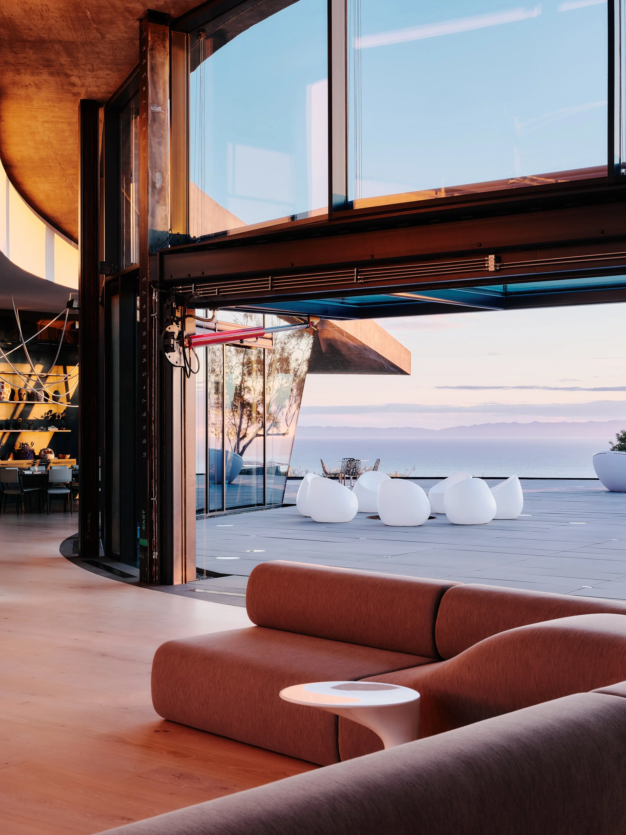 Modern interior living space with a view of the ocean. There are white bubble-shaped chairs outside on a deck, and a large window or sliding door leading to the outdoor area. The interior features warm wood tones and curved furniture, creating a contemporary and inviting ambiance.