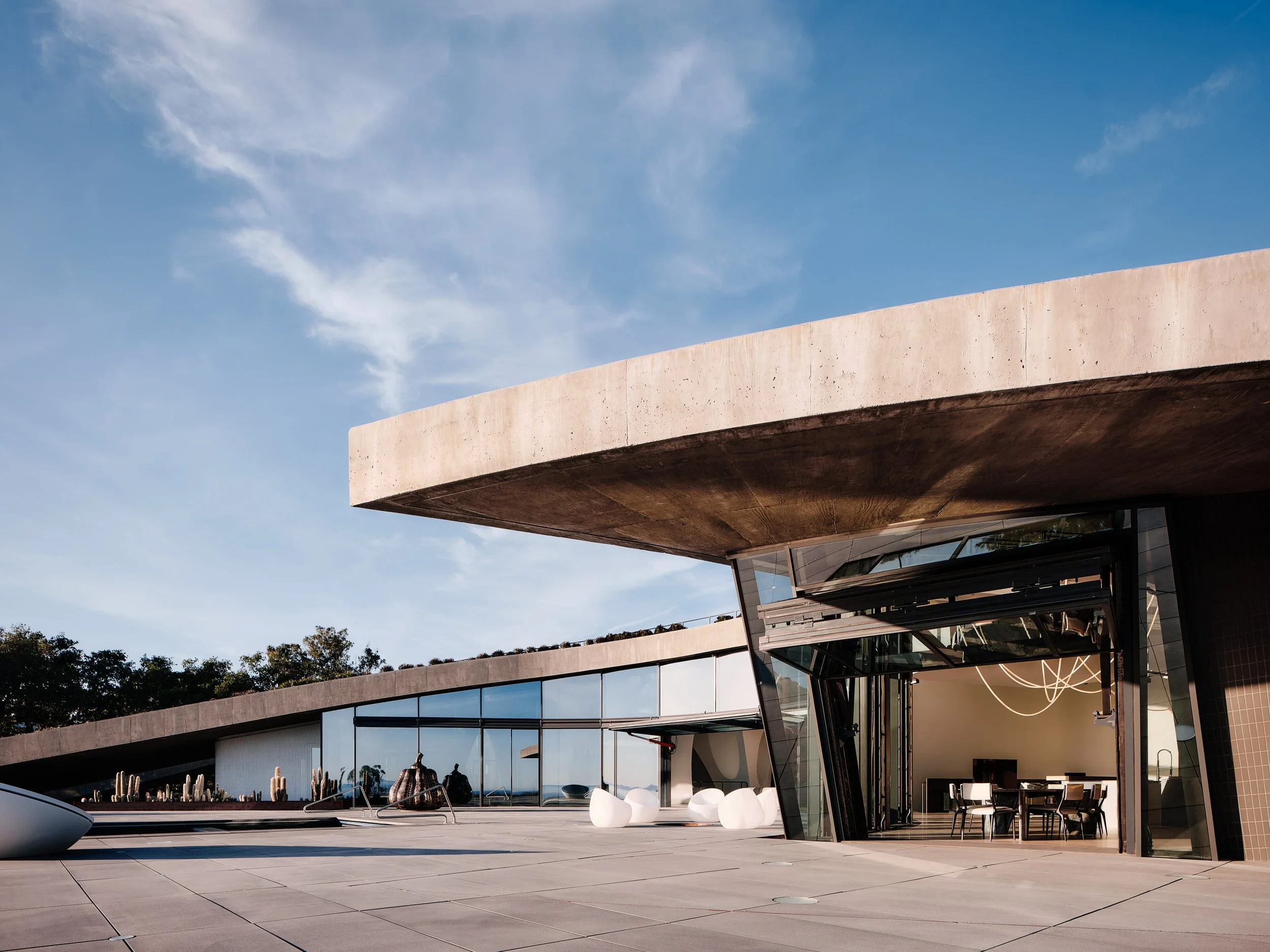 Modern building with large glass windows and an overhanging concrete structure, with a clear blue sky and some trees in the background.