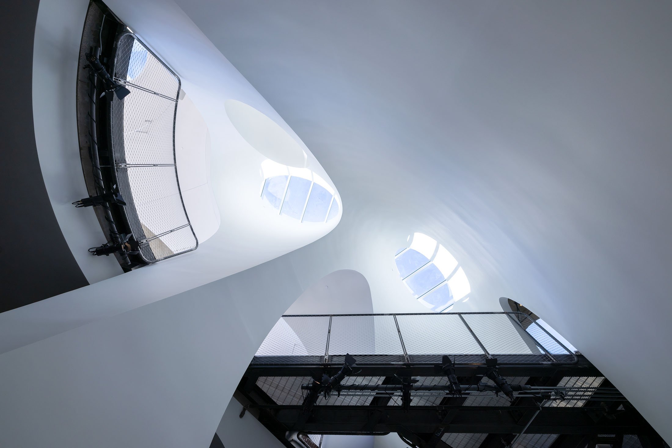 Interior of a modern building with white curved walls, large round windows, a metal staircase with a black railing, and some lighting equipment at the bottom.